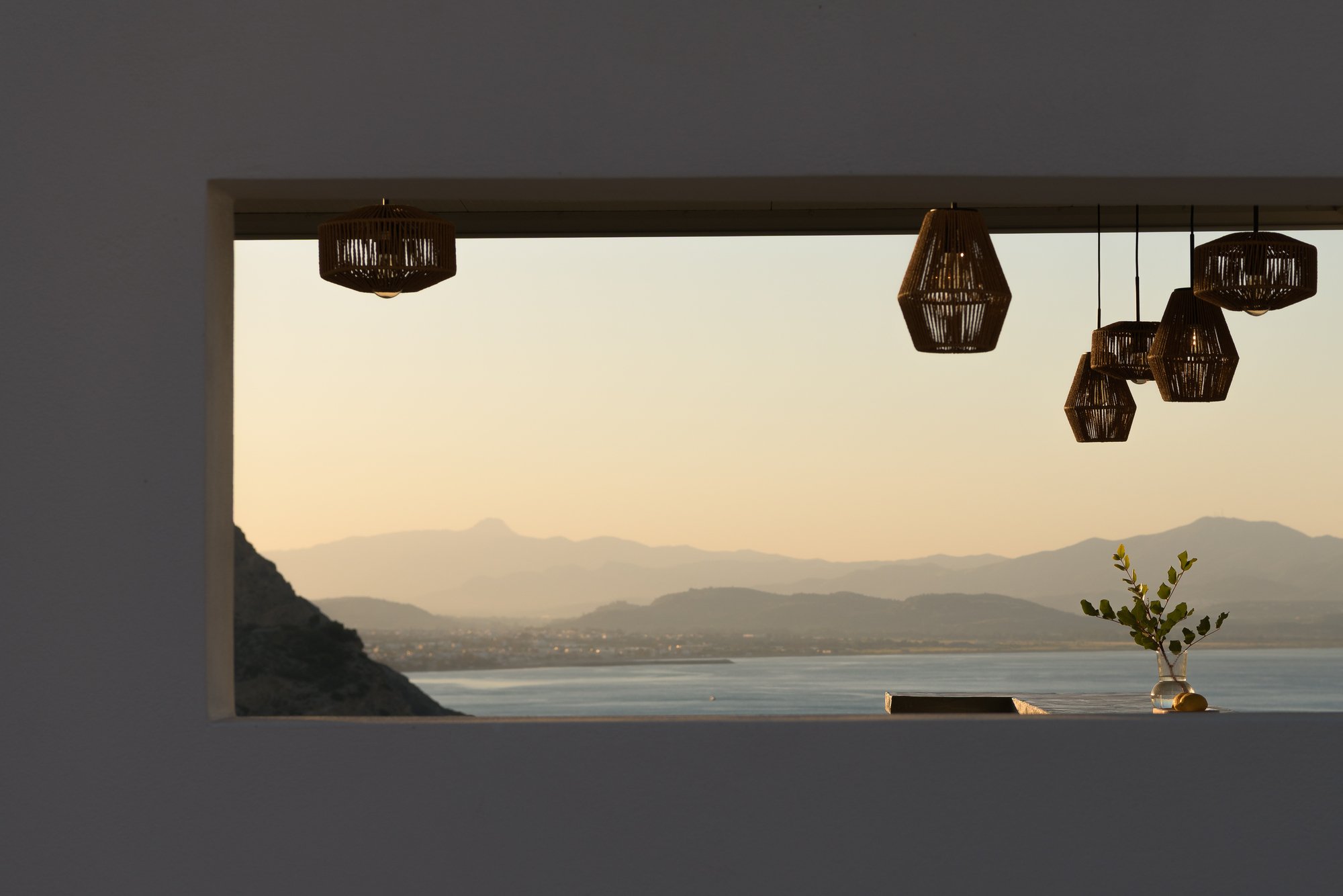 View through a large window opening at Galini Breeze in Agia Galini showing a scenic landscape with mountains, sea, and a sunset sky. Hanging wicker pendant lights and a glass vase with a small plant are visible in the foreground.