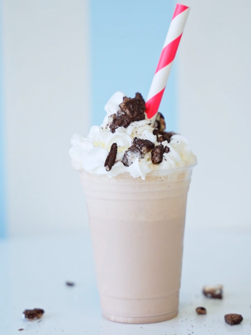 Chocolate milkshake topped with whipped cream and chocolate cookie crumbles, with a red and white striped straw, on a white surface with scattered cookie crumbles.