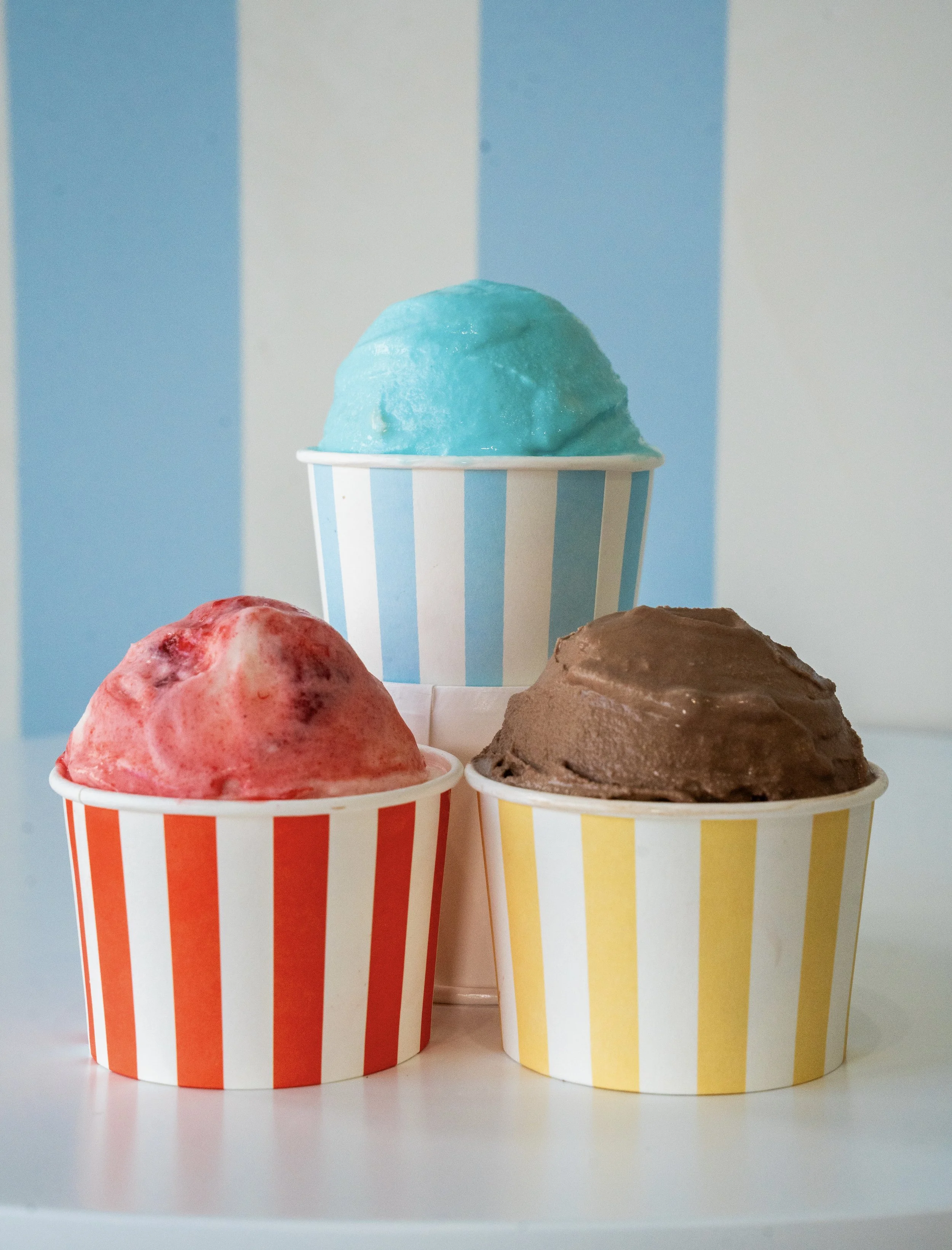Three cups of ice cream, each with a different flavor and color, in striped paper cups against a background with vertical blue and white stripes.