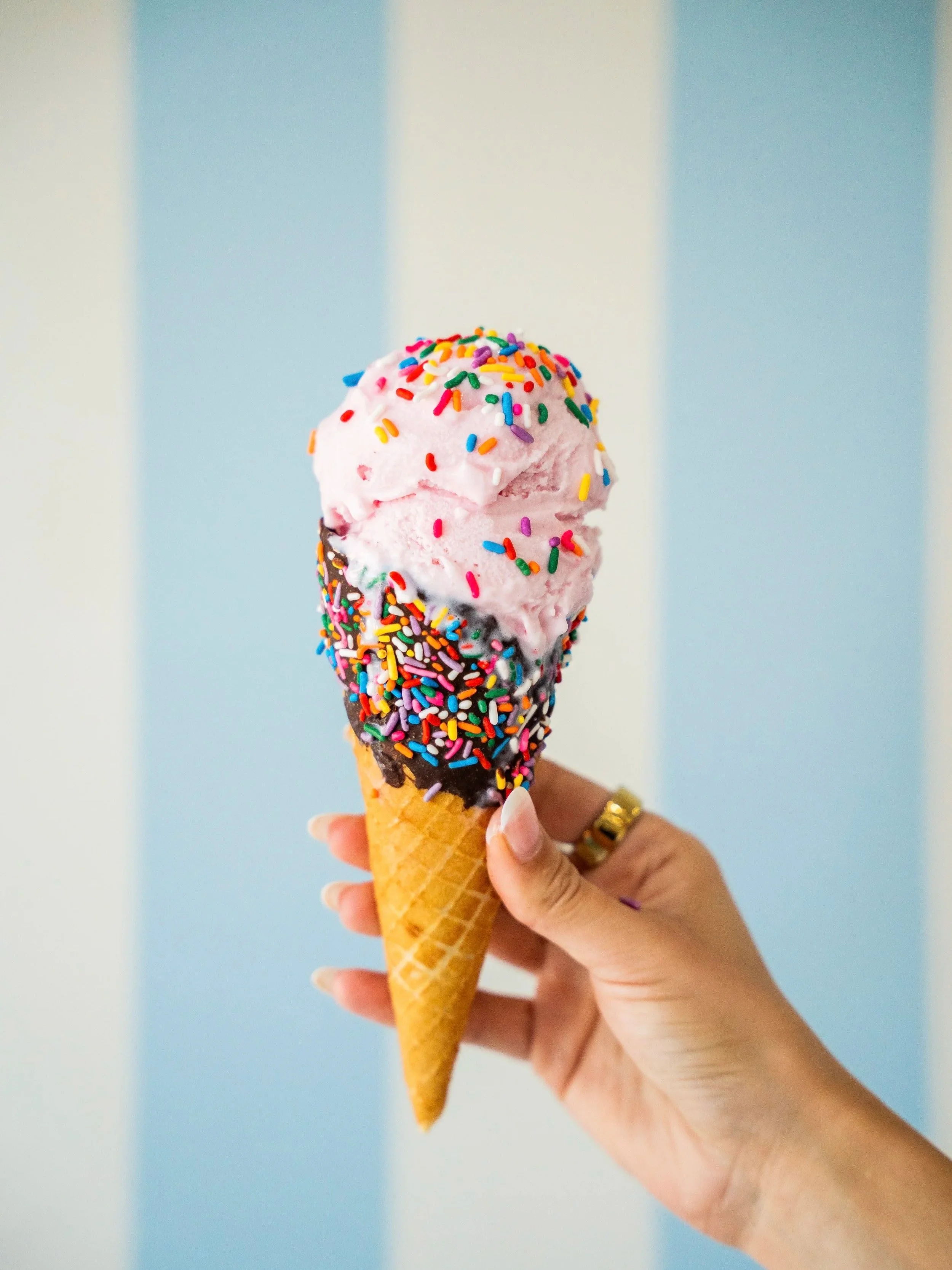 A hand holding a two-scoop ice cream cone with rainbow sprinkles against a vertical striped blue and white wall.