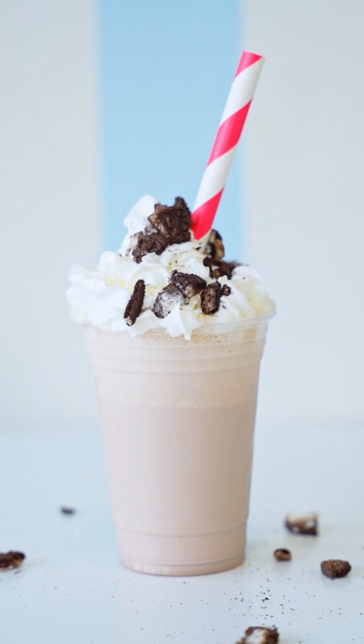 An oreo shake in a plastic cup with whip cream and Oreos on top on a table with a red and white striped straw coming out of it.