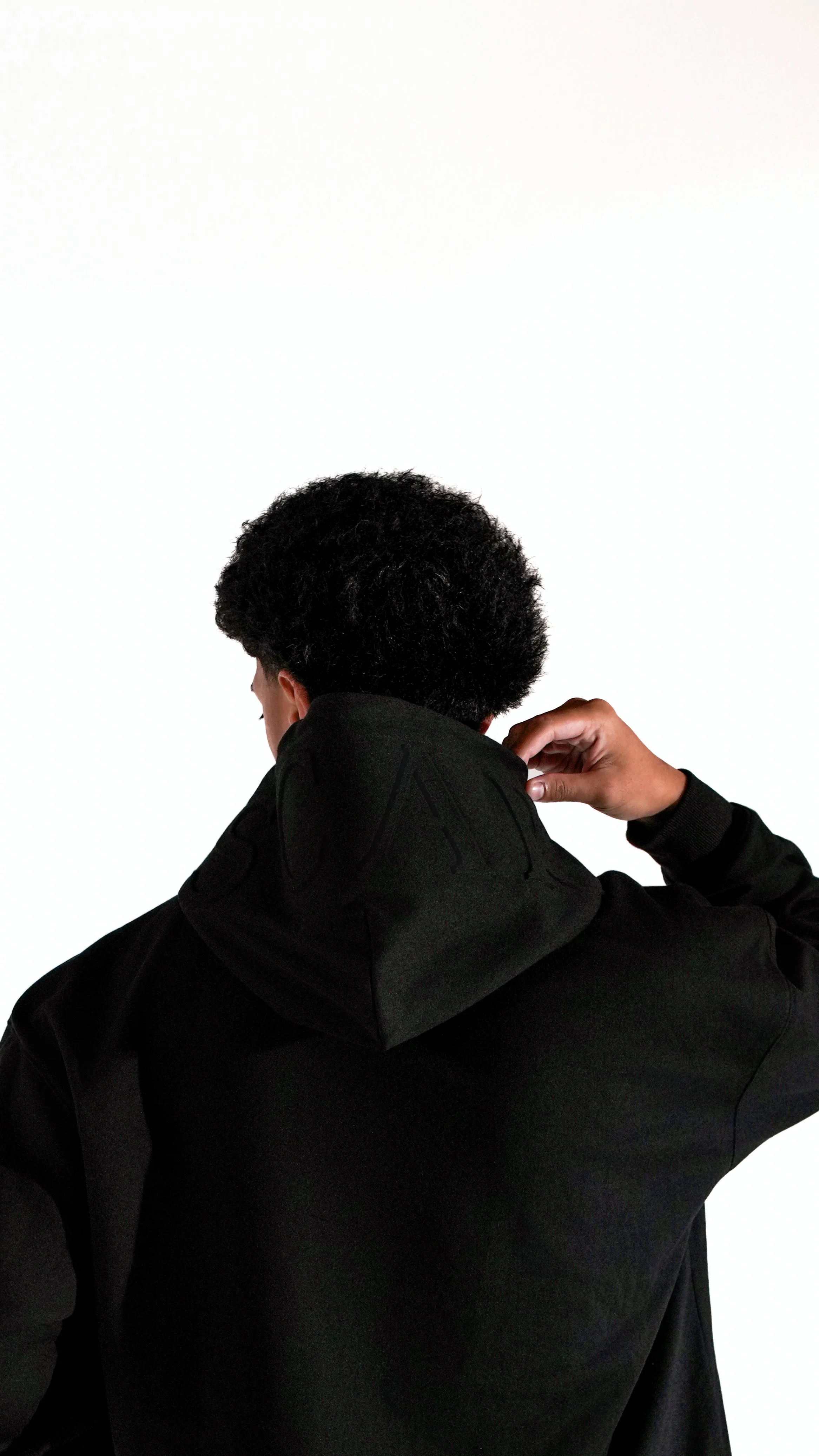 Rear view of a person with curly hair, wearing a black hoodie carefully embossed, adjusting the collar of the hoodie with one hand, against a plain white background.