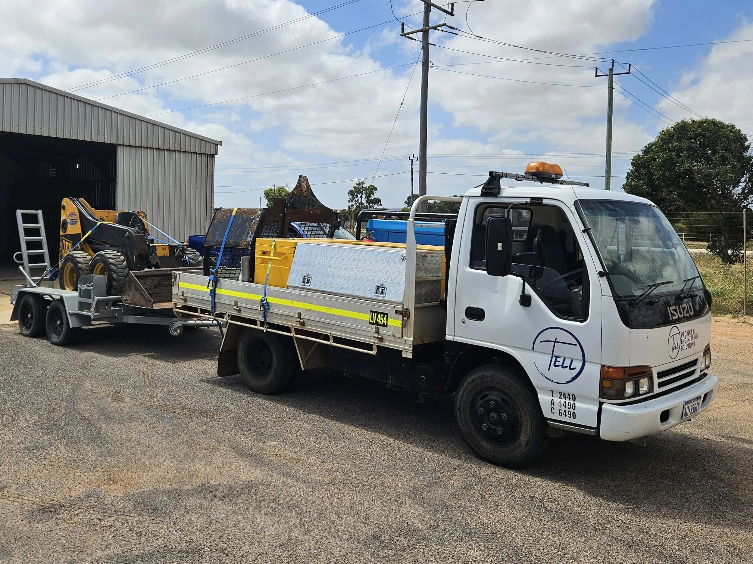 A white flatbed truck with a yellow flashing light on top, carrying equipment and construction materials, parked in front of a metal building with graffiti on a partly cloudy day.
