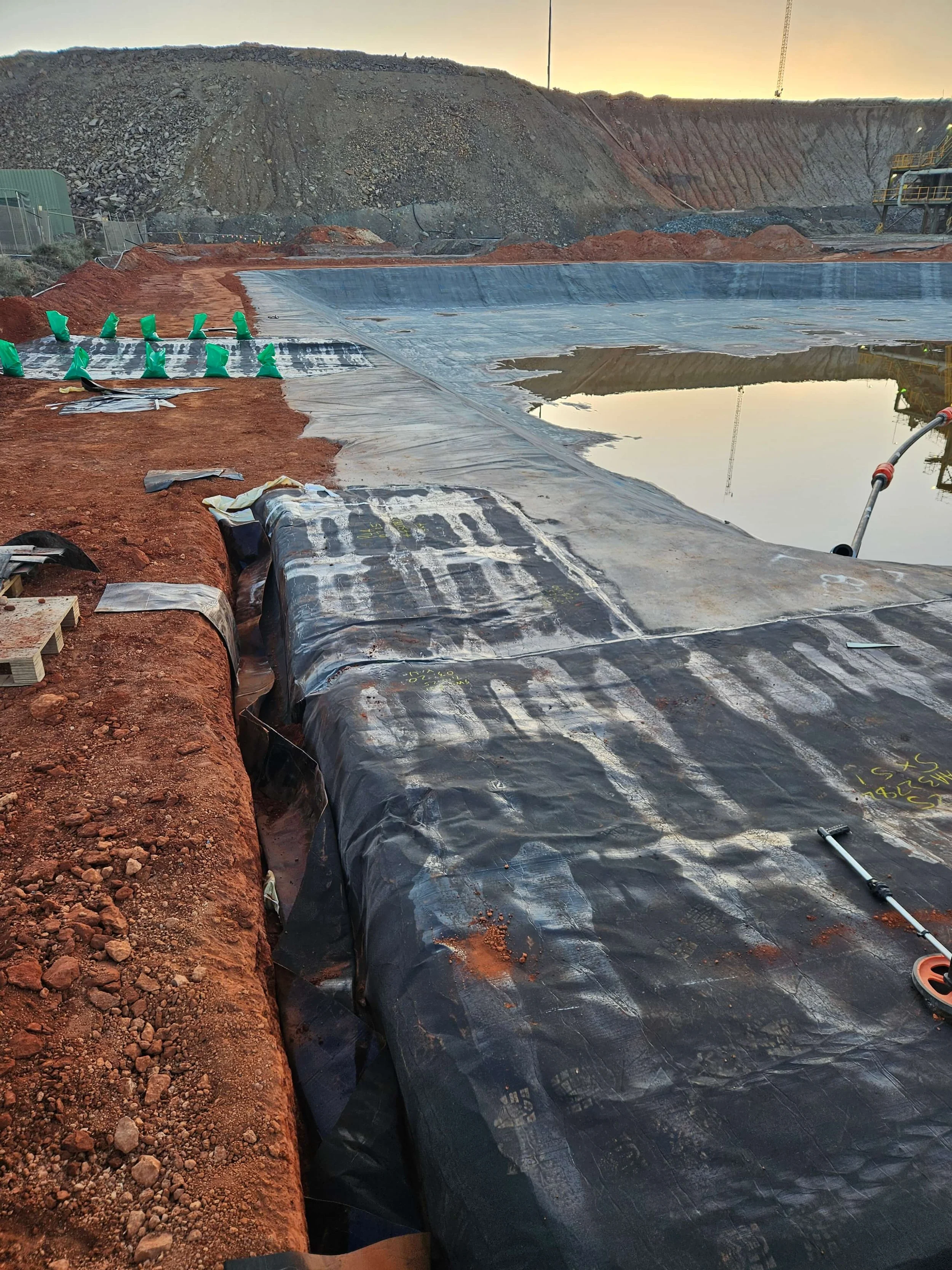 Construction site with a large excavated area covered with black plastic sheeting, red dirt, and a water pond, with rocky hills and a crane in the background during sunset.