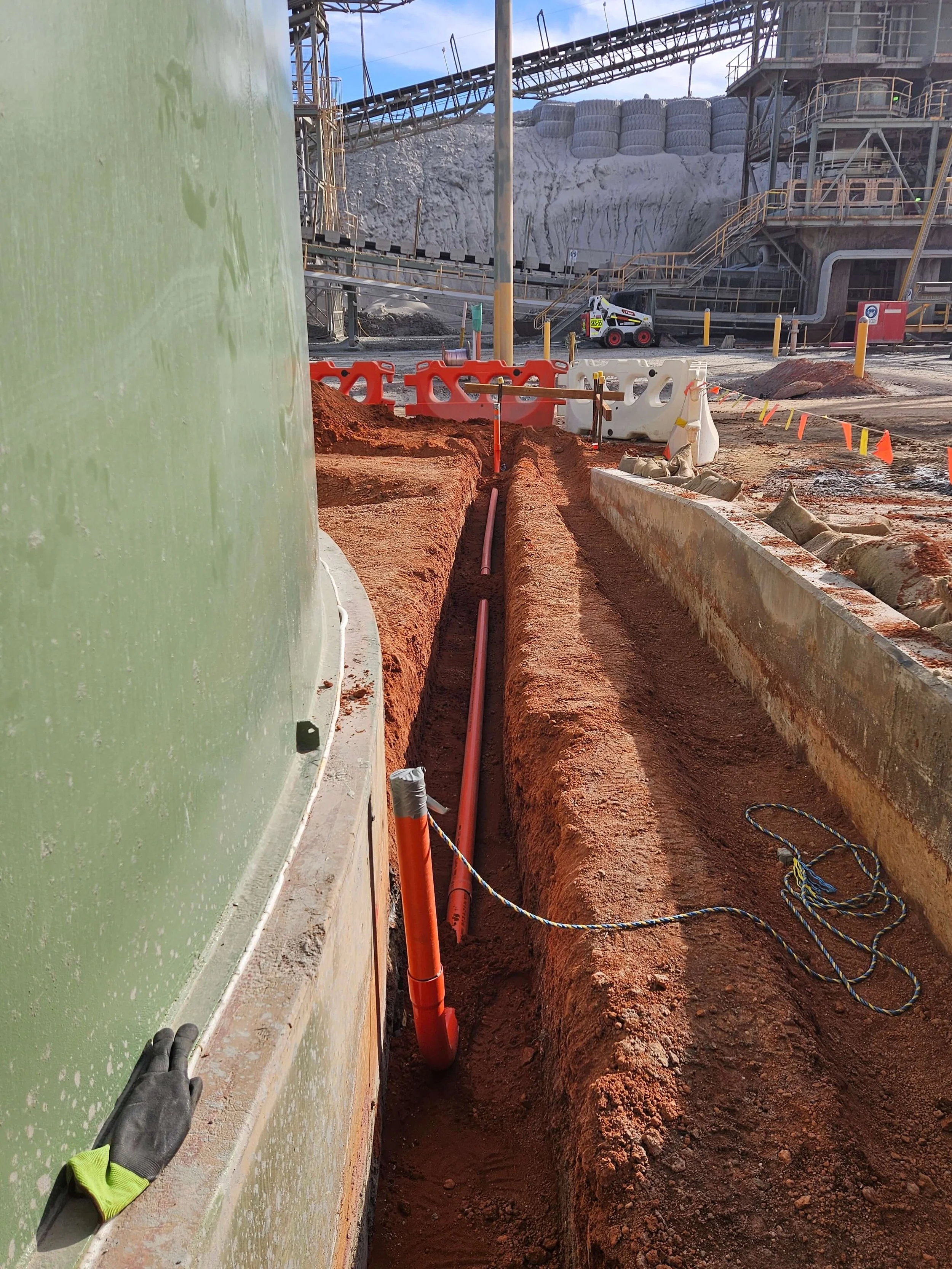 Construction site with a deep trench for underground piping or cables, orange pipes in the trench, safety barriers, heavy industrial equipment in the background, and a worker's gloved hand visible on the left side.