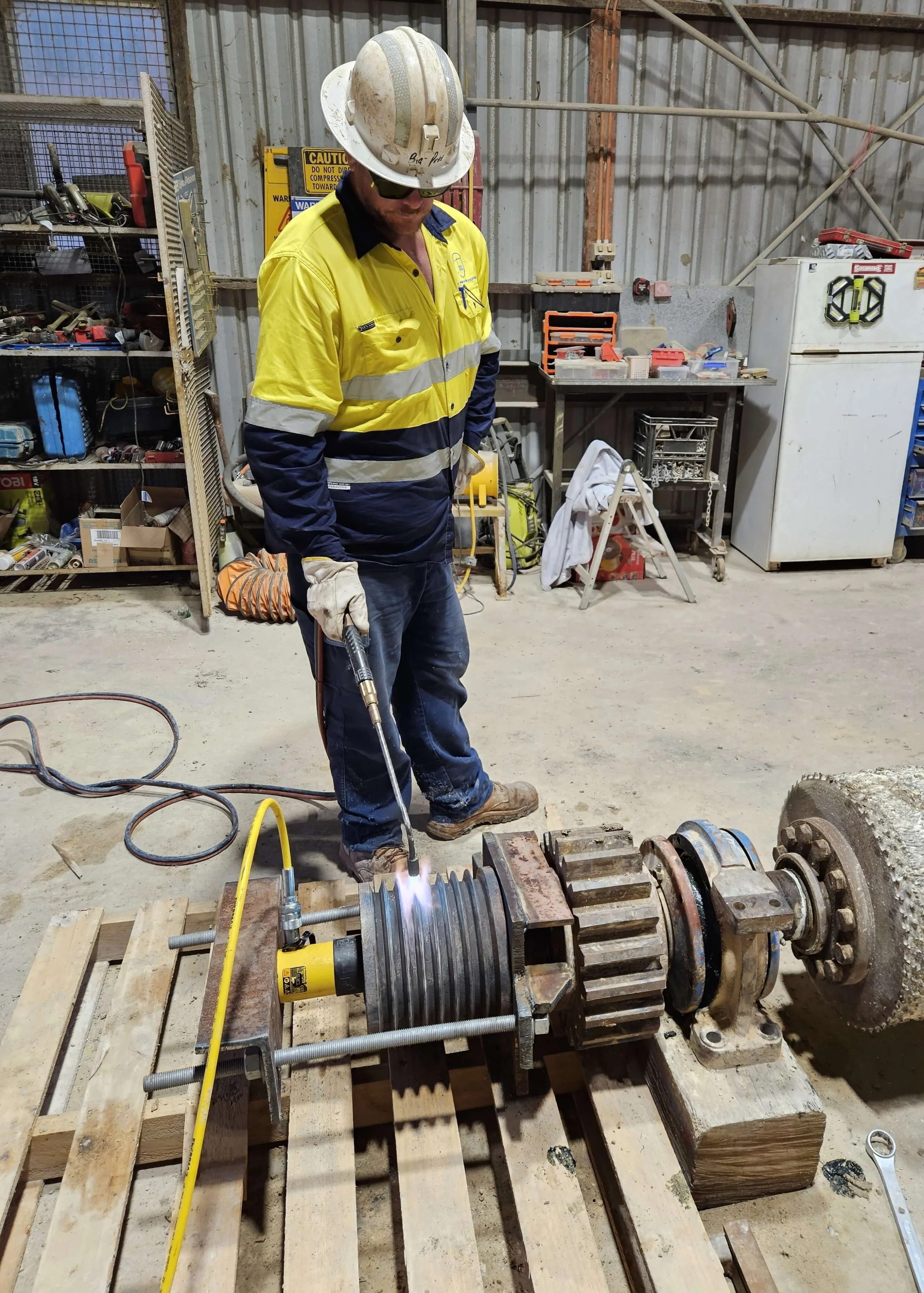A worker in a yellow safety jacket and white hard hat is welding a large metal gear assembly in an industrial workshop. The gear is clamped to a wooden platform, with various tools and equipment visible in the background.