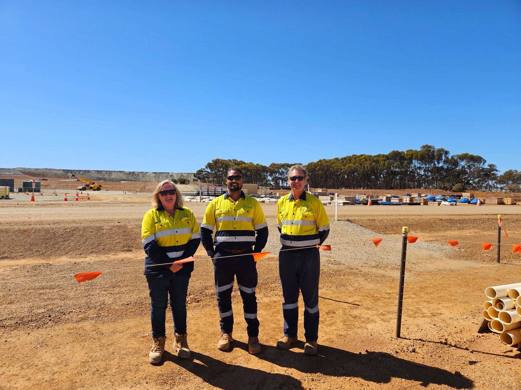 Three construction workers in yellow safety jackets standing on a construction site with clear blue sky and trees in the background.