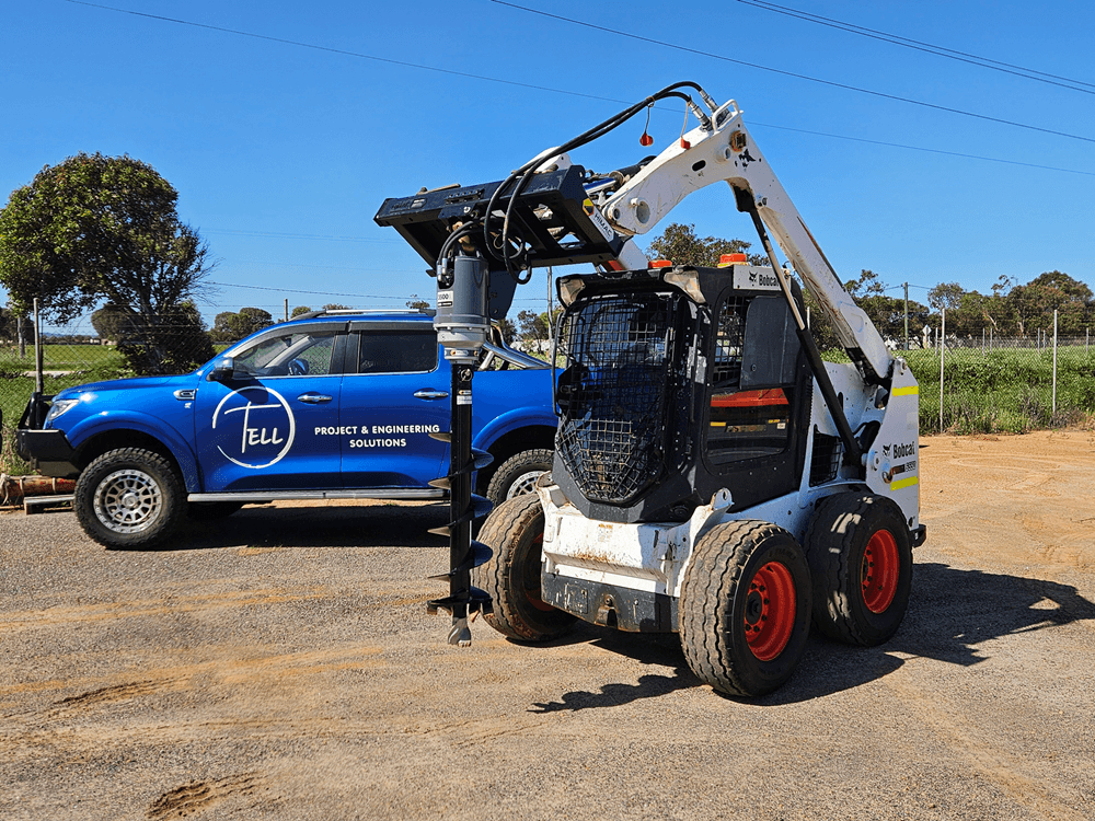 A skid-steer loader parked next to a blue project vehicle with the logo 'TELL Project & Engineering Solutions' on the side, on a dirt lot with trees and power lines in the background.