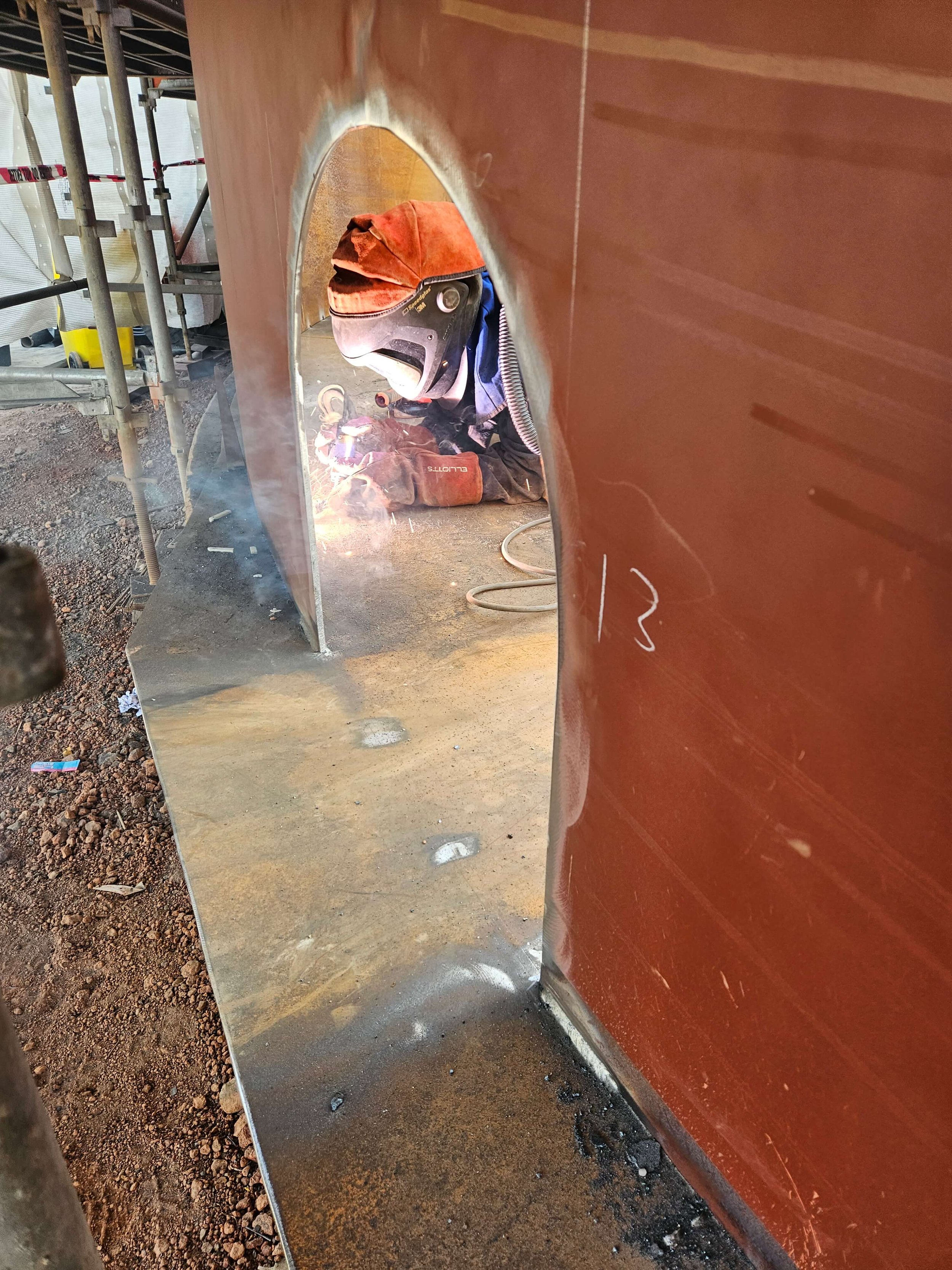 A welder in safety gear welding a large metal structure at a construction site, with sparks visible.