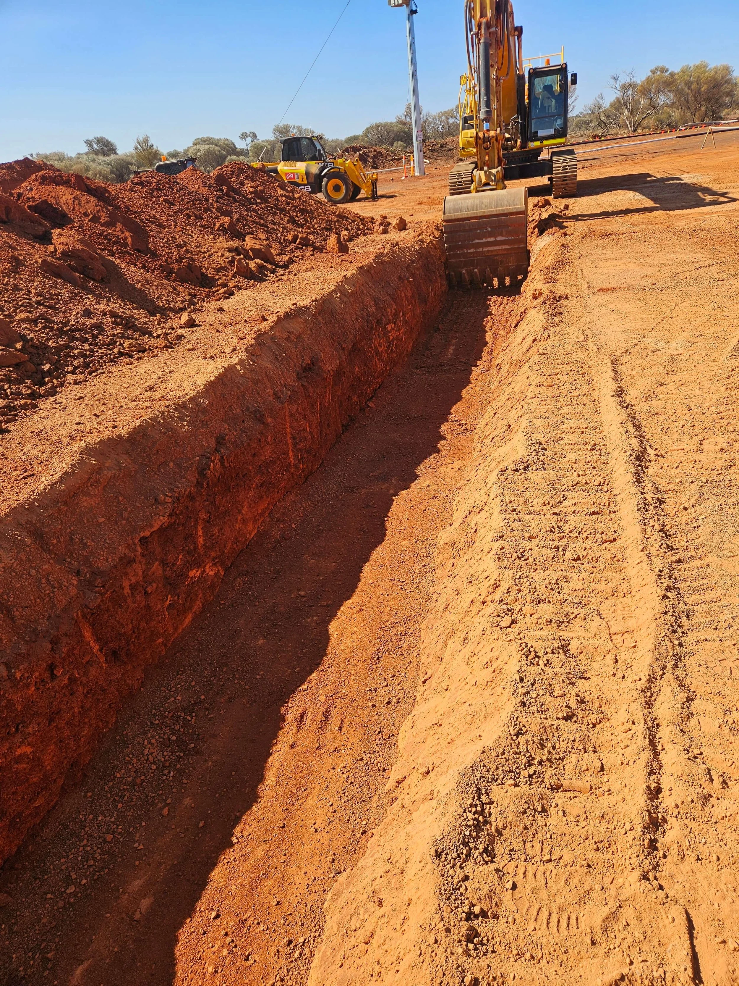 Construction site with excavator and bulldozer working on a dirt road, clearing and leveling the ground under a clear blue sky.