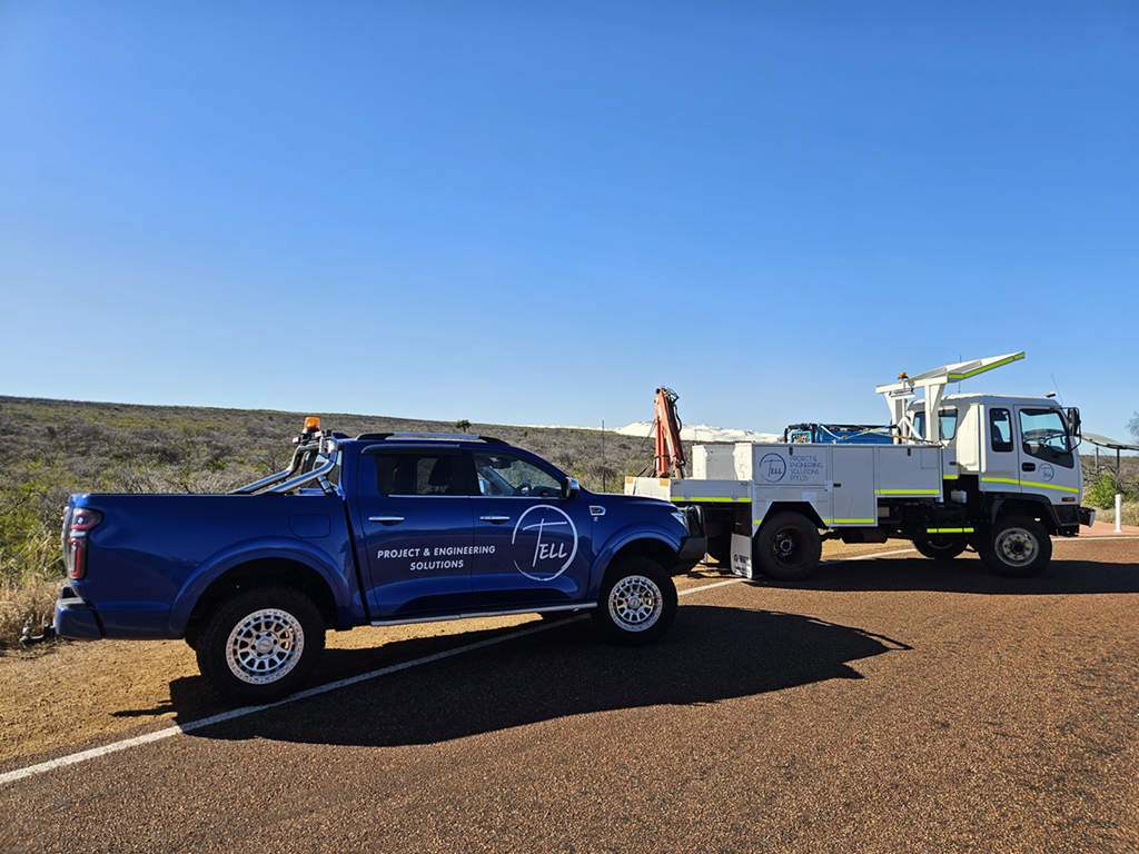 A blue project and engineering solutions pickup truck parked beside a white utility truck on an open road with a clear blue sky and arid landscape in the background.