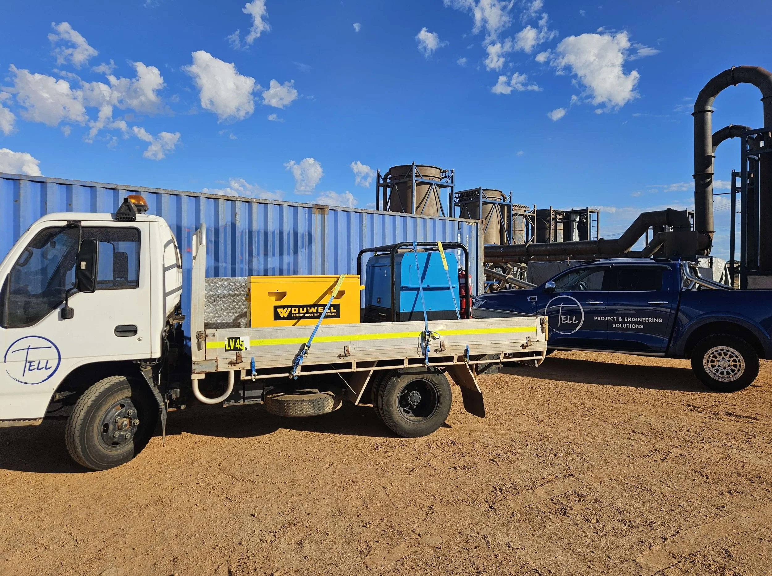 A white utility truck with a flatbed carrying yellow, blue, and black equipment, parked on dirt ground under a blue sky with scattered white clouds. The truck has a cab with a side mirror and a warning light on top.