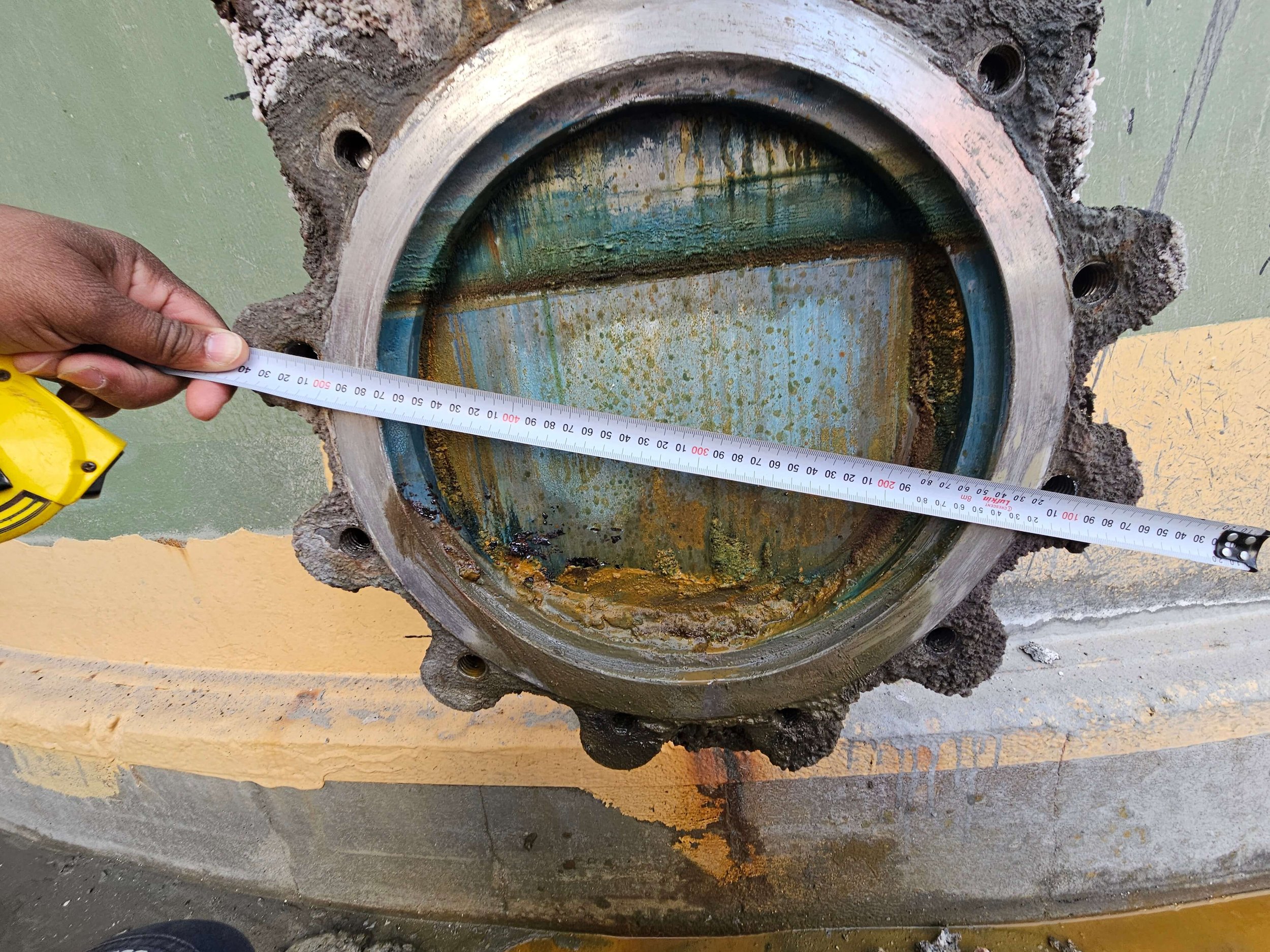 Close-up of a large, rusty, and corroded metal pipe flange with a person holding a measuring tape across its diameter. The flange has four bolt holes and signs of corrosion and rust inside and around it. The background includes a green wall and some 