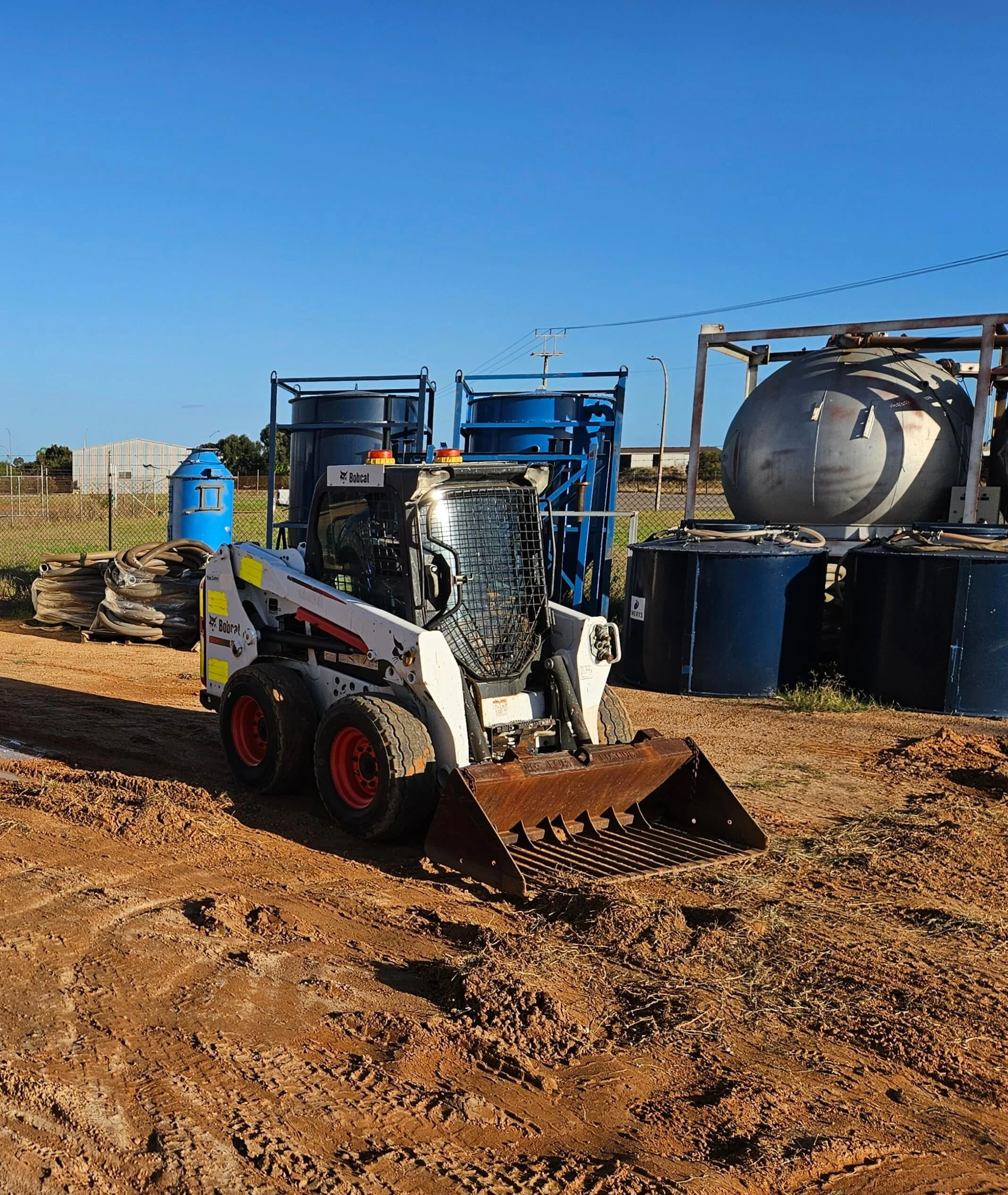 Construction site with dirt ground, a small skid-steer loader, large blue tanks, and a large rusted metal tank, under a clear blue sky.