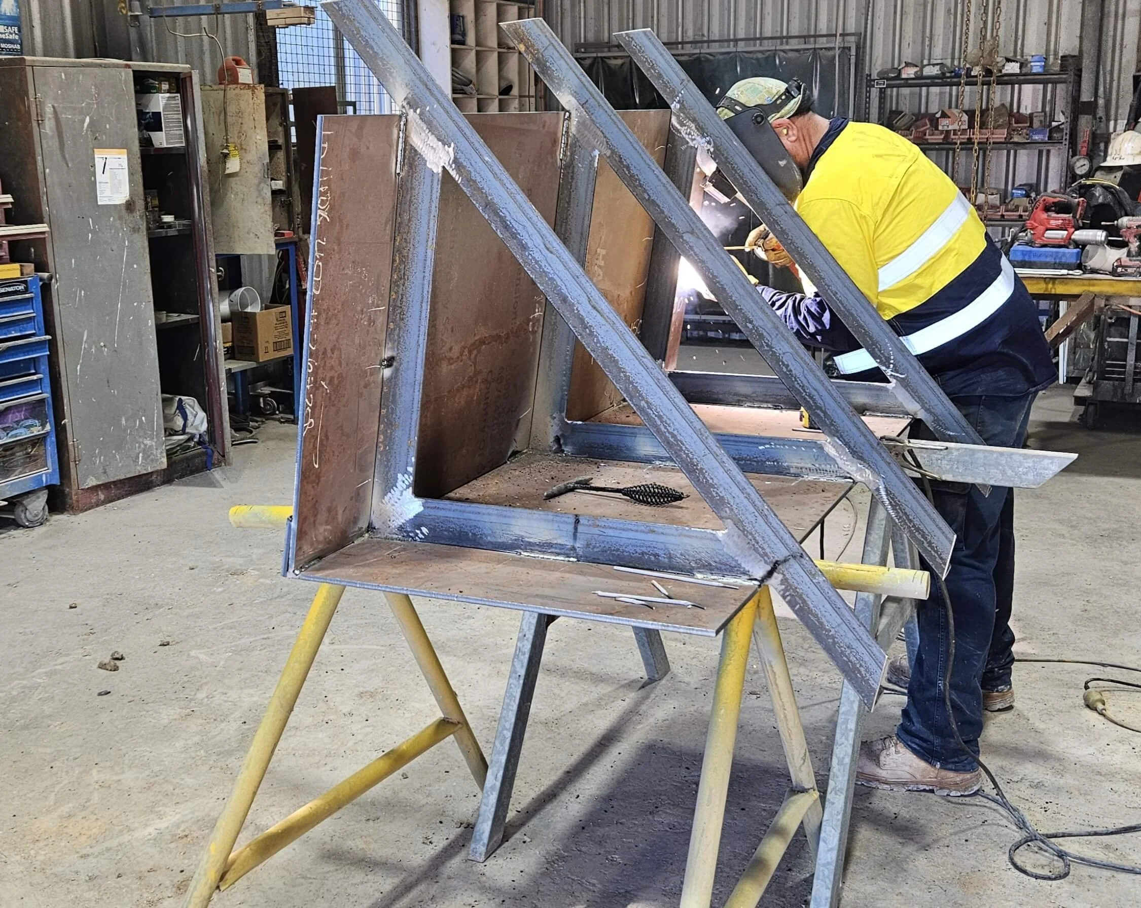A worker welding metal framework inside a workshop, wearing safety gear including a helmet and reflective vest, with various tools and materials around.