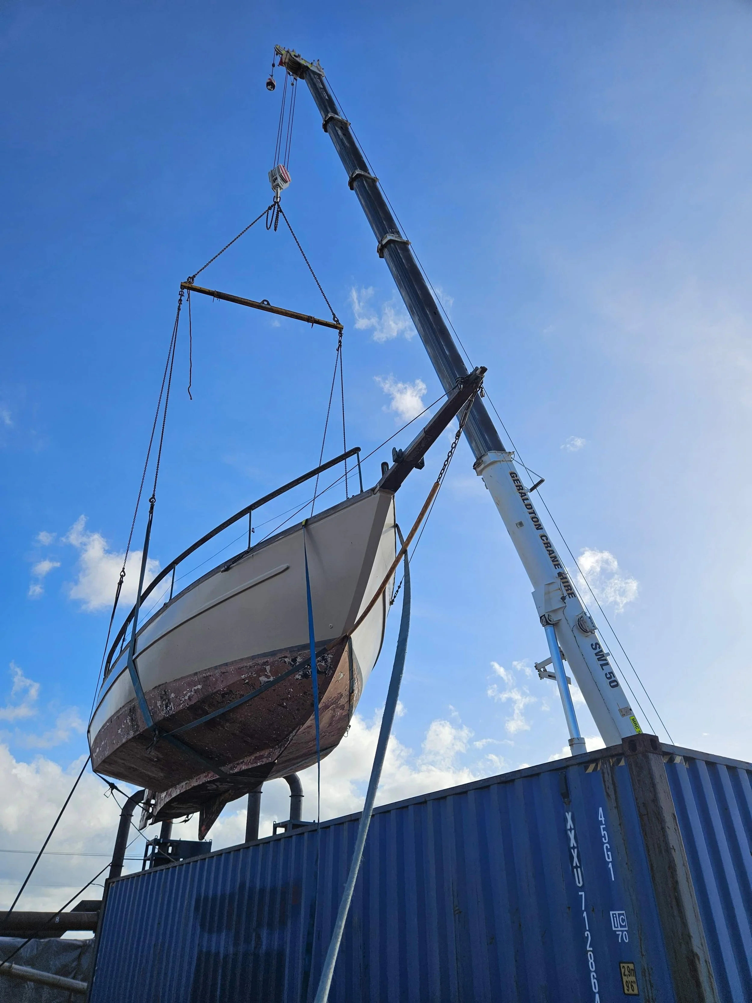 A boat is being lifted for repair by a crane at a dock, with a blue shipping container below and a partly cloudy sky in the background.