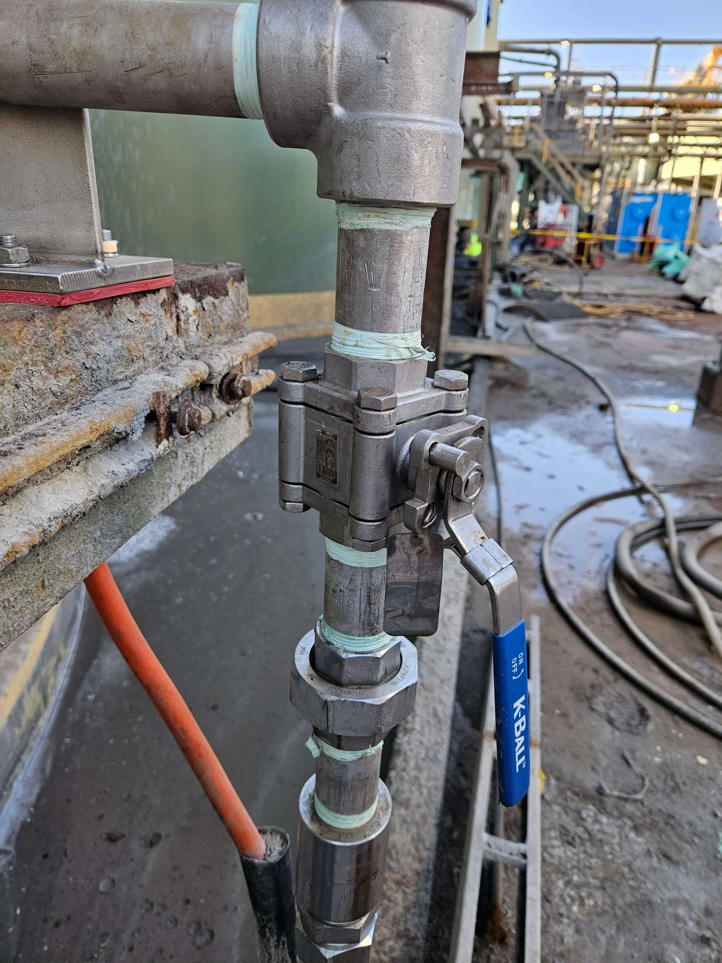 Close-up view of industrial piping with valves, fittings, and insulation, set in an industrial environment with other pipes and equipment in the background.