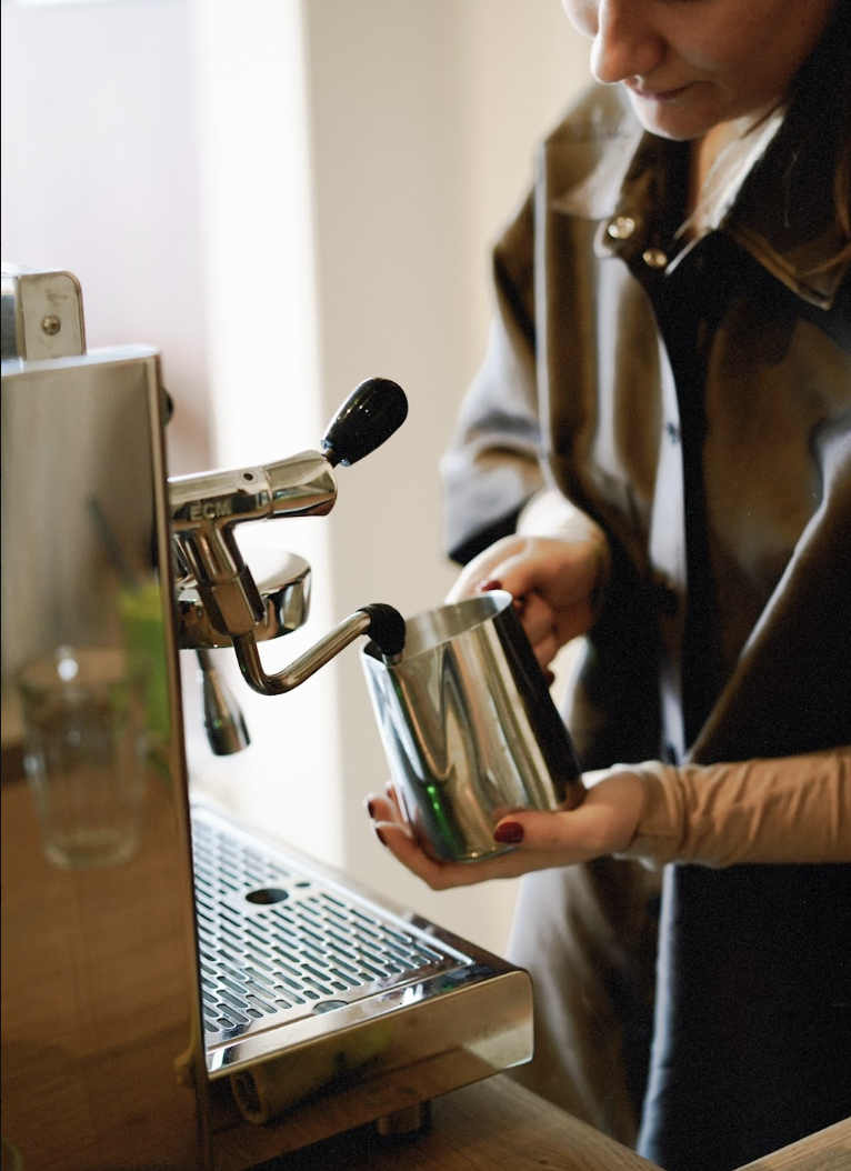 Person using an espresso machine, holding a metallic milk frothing pitcher, preparing coffee.