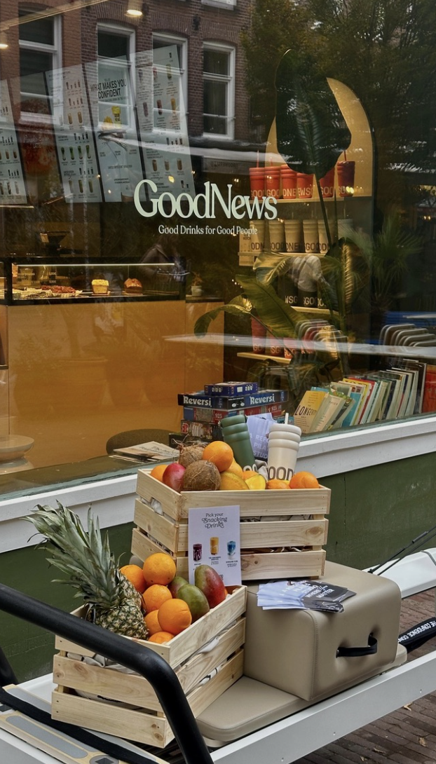 Outside display of a grocery store with wooden crates of fruits including pineapple, oranges, apples, and coconuts, along with cups and promotional flyers on a cart in front of the store window.