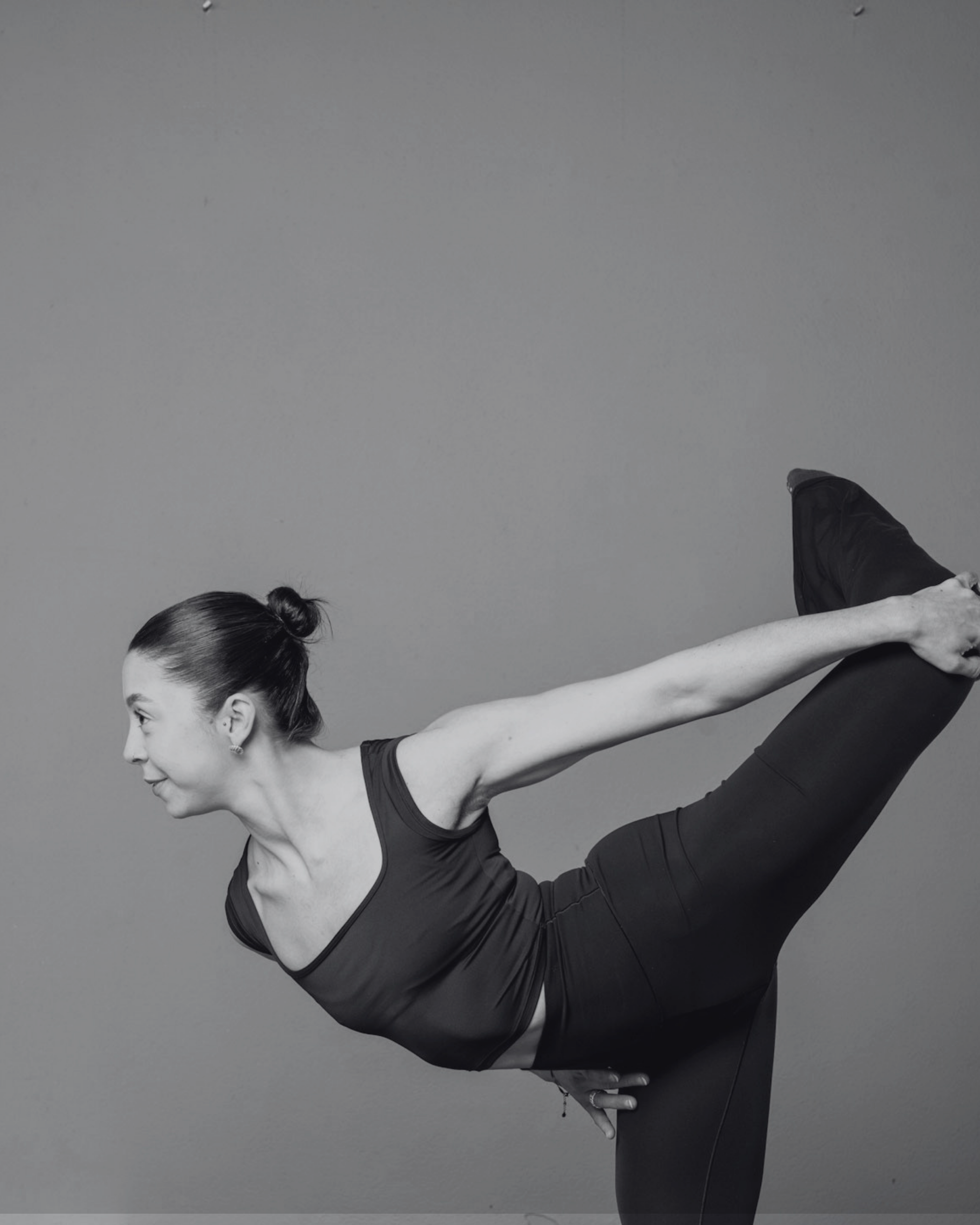 A woman with dark hair tied in a bun, wearing a black workout top, performing a yoga pose against a plain gray wall.