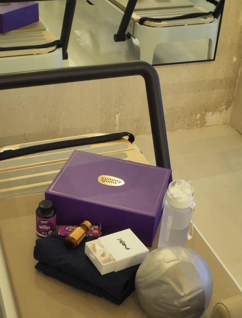 A mirror reflecting a purple box, a water bottle, a small bottle, a pair of black gloves, a box of tissues, and a round white object on a beige surface.
