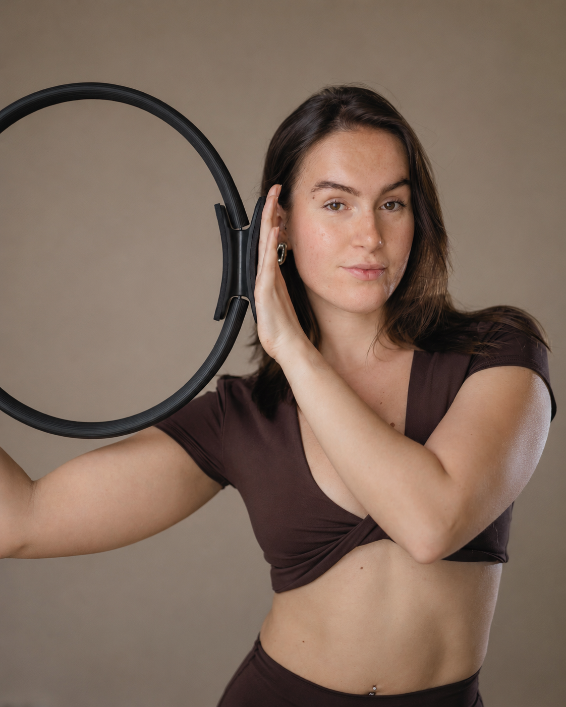 A young woman with long brown hair, wearing a brown crop top, holding a black circular object, and touching her ear against it.