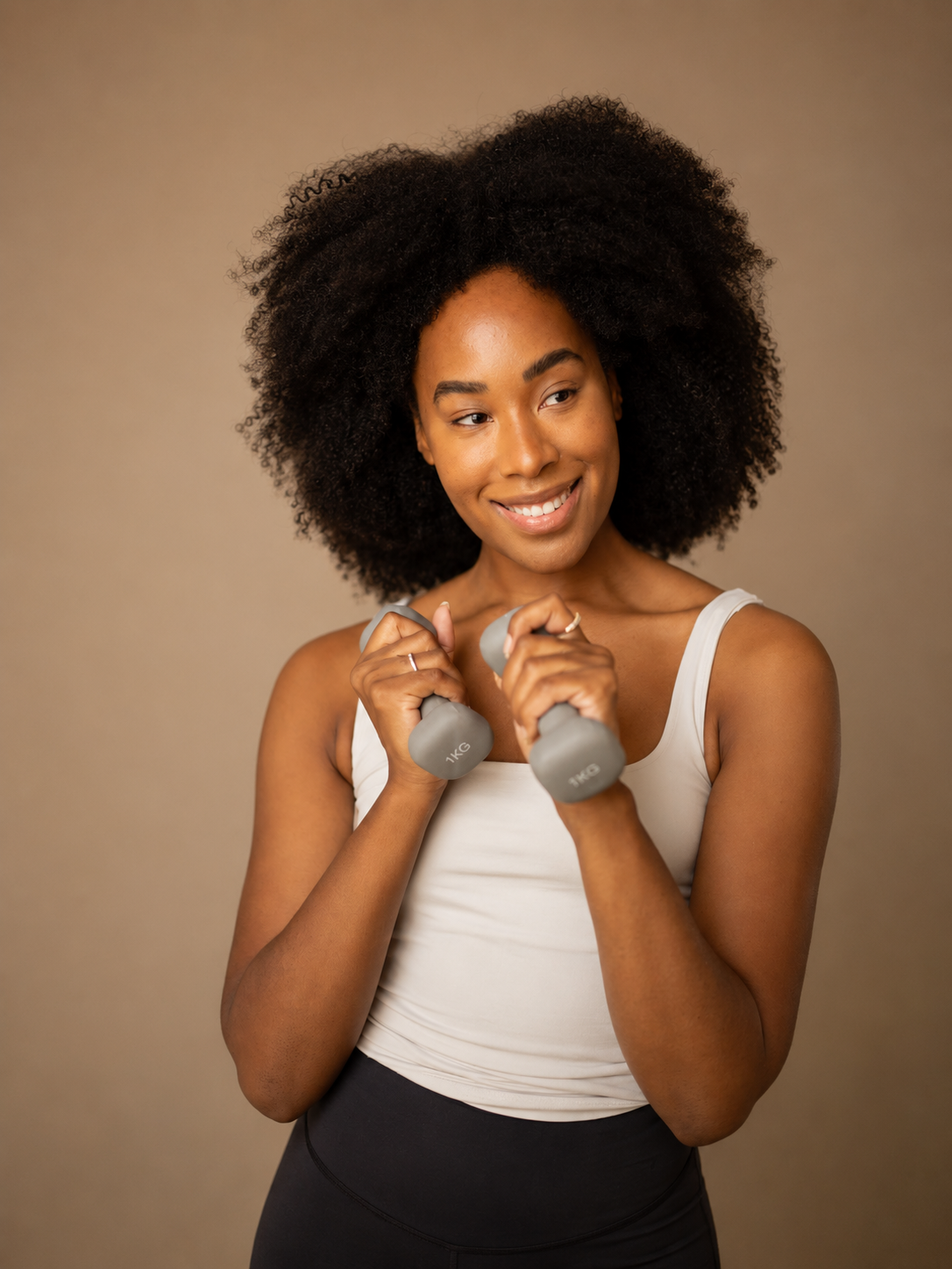 A woman with curly black hair holding two gray dumbbells, smiling and looking to her left.