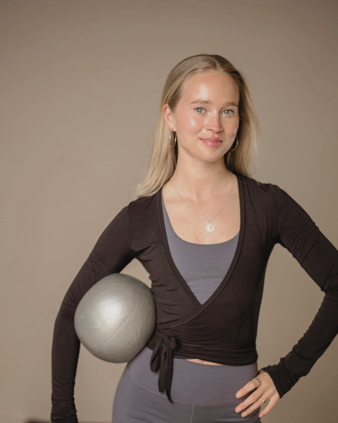 Woman with blonde hair, wearing a dark wrap-top, grey workout shirt, and grey leggings, holding a silver exercise ball against her side, posing with one hand on her hip against a beige background.