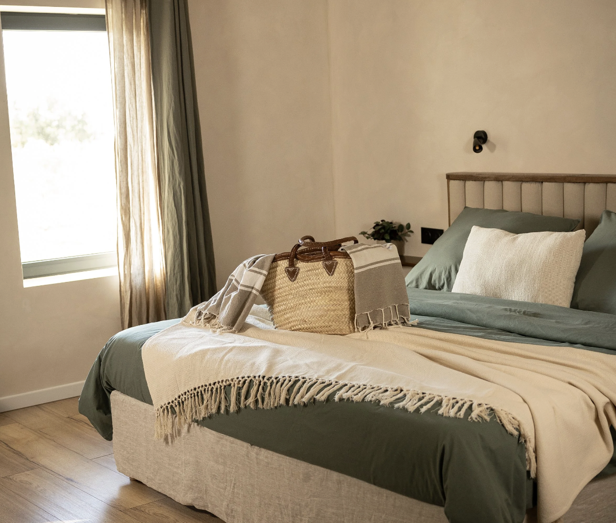 A neatly made bedroom with a bed covered in green and cream bedding, a straw bag with a beige towel on top sitting on the bed, and a window with beige curtains letting in natural light.
