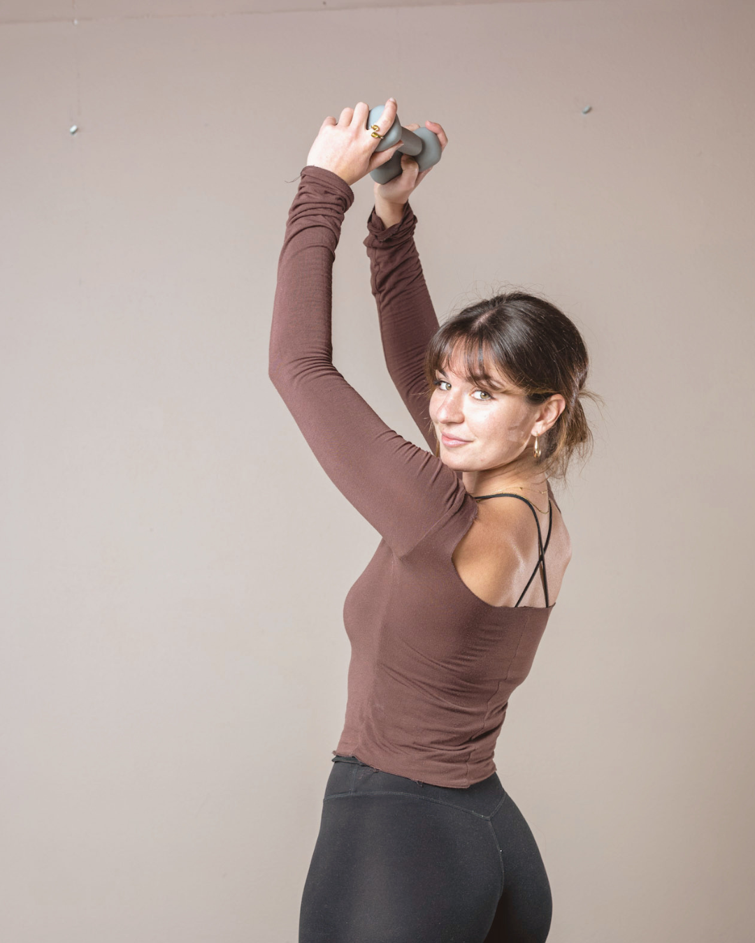 A woman with brown hair in a ponytail, wearing a brown long-sleeve top and black leggings, holds a gray dumbbell overhead and looks at the camera with a slight smile.