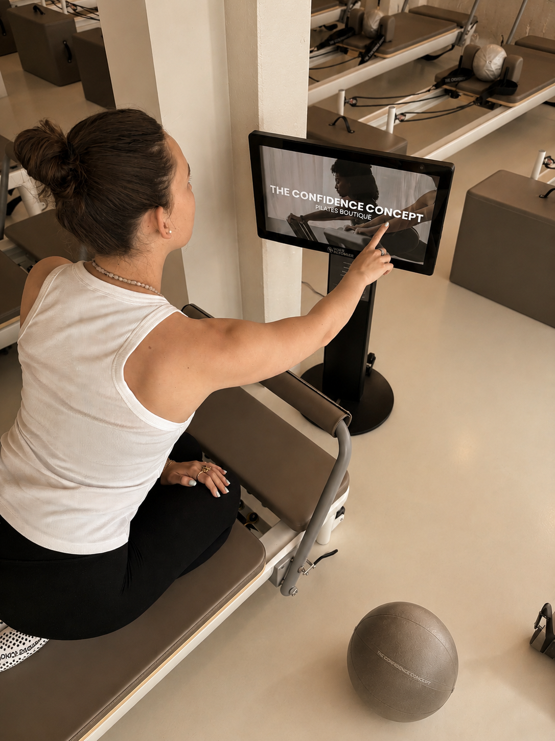 Woman sitting on a Pilates reformer machine at a Pilates boutique, pointing at an exercise screen that reads 'The Confidence Concept Pilates Boutique', with Pilates equipment and a exercise ball nearby.