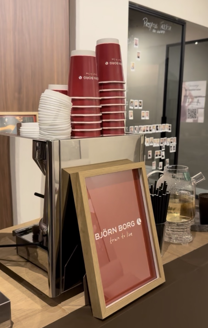 Coffee shop counter with red cups stacked, white cups, a framed sign reading "BJÖRN BORG train to live," and a glass pitcher of water with a glass and black straws.