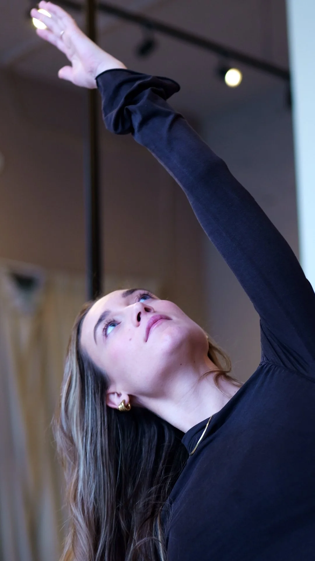 A woman with long hair and earrings is reaching up with her arm extended, looking upward.