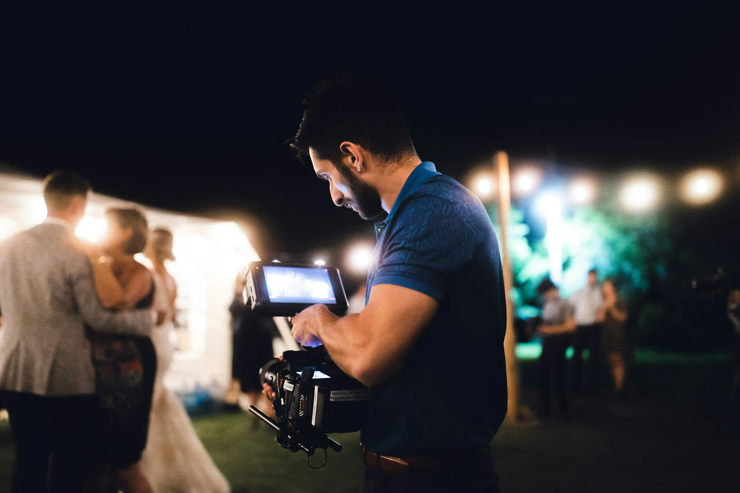 A man with dark hair and a beard operating a camera at an outdoor event at night, with blurred people dancing and colorful lights in the background.