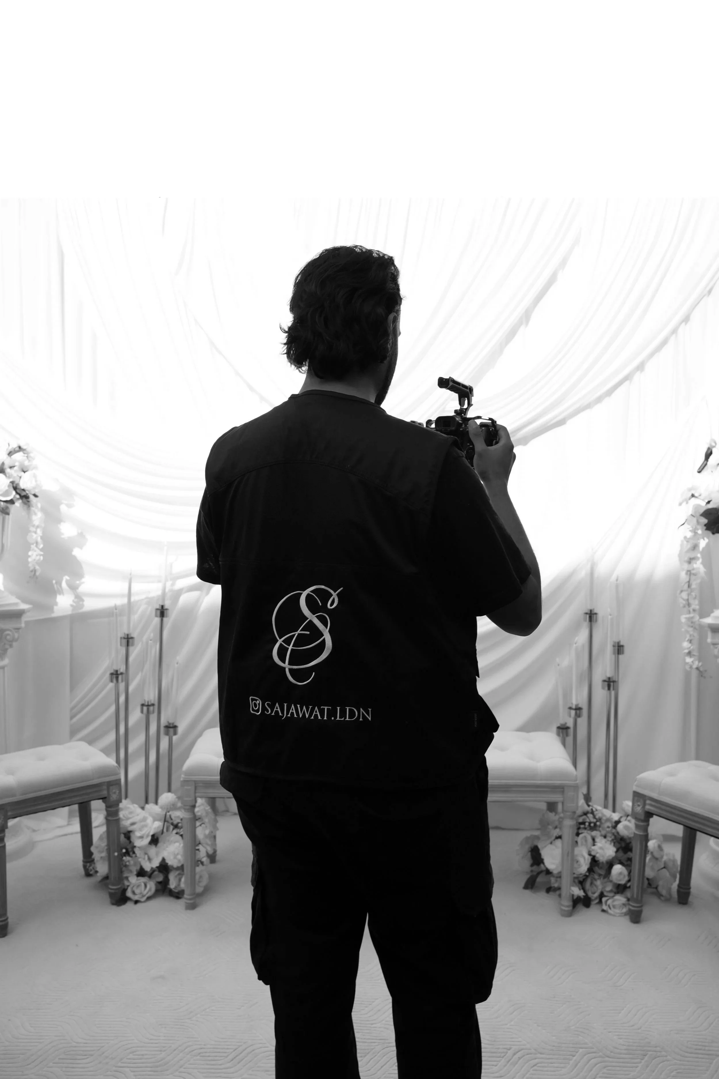 A man with long dark hair takes a photo in a black-and-white room decorated for a wedding with chairs, flower arrangements, and drapery.