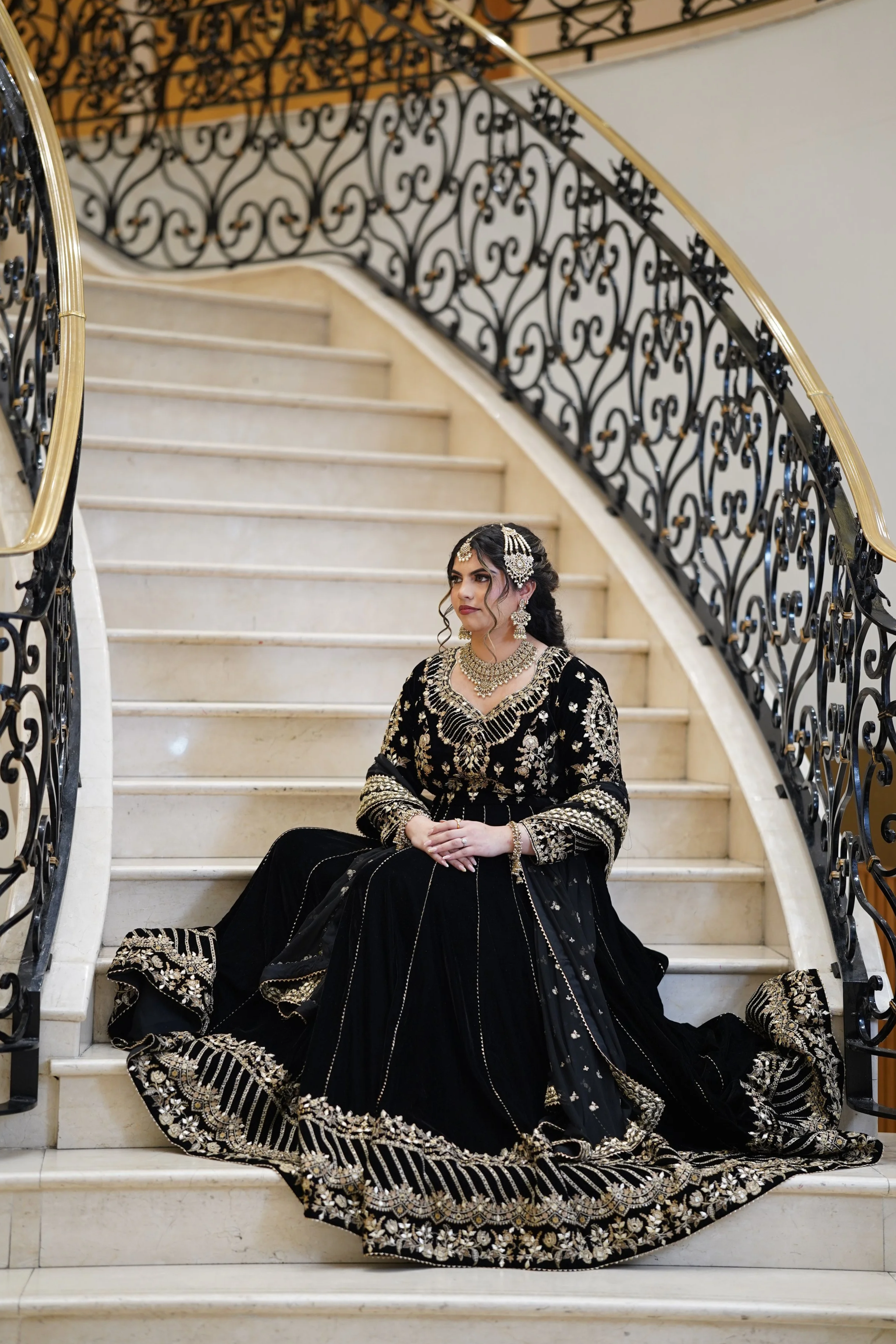 Woman dressed in traditional black and gold bridal attire sitting on a staircase with ornate black and gold railing.