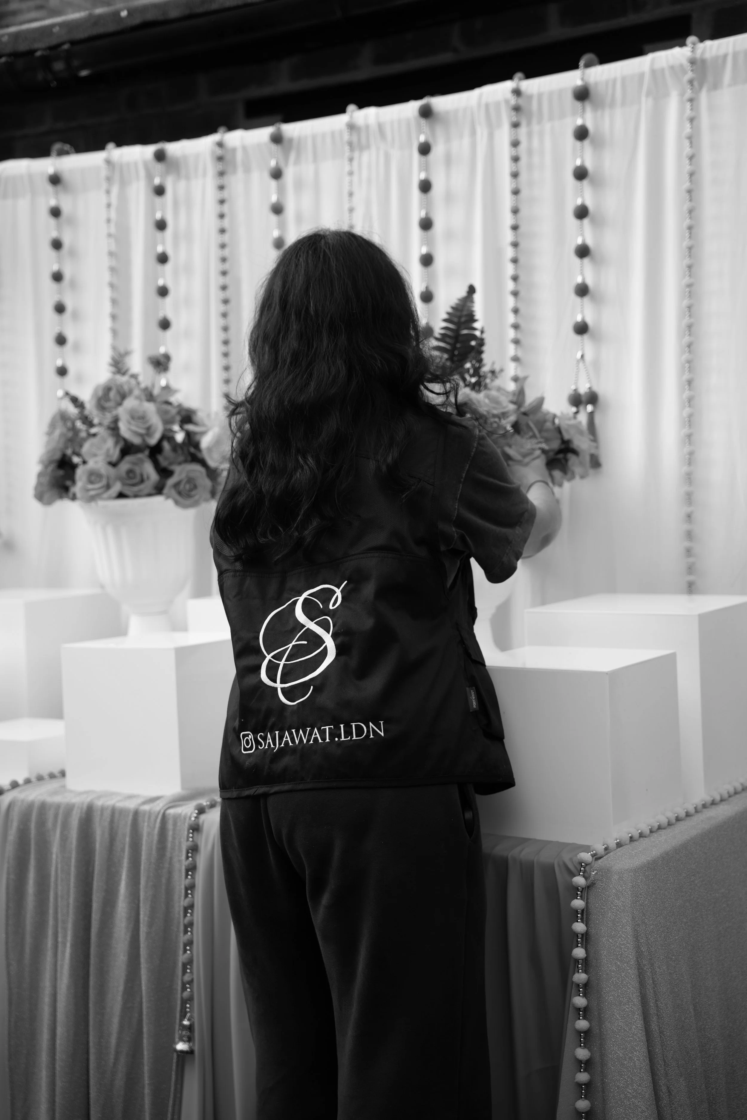 Person with long dark hair standing in front of a white curtain decorated with hanging beads and flowers, holding a bouquet of roses.