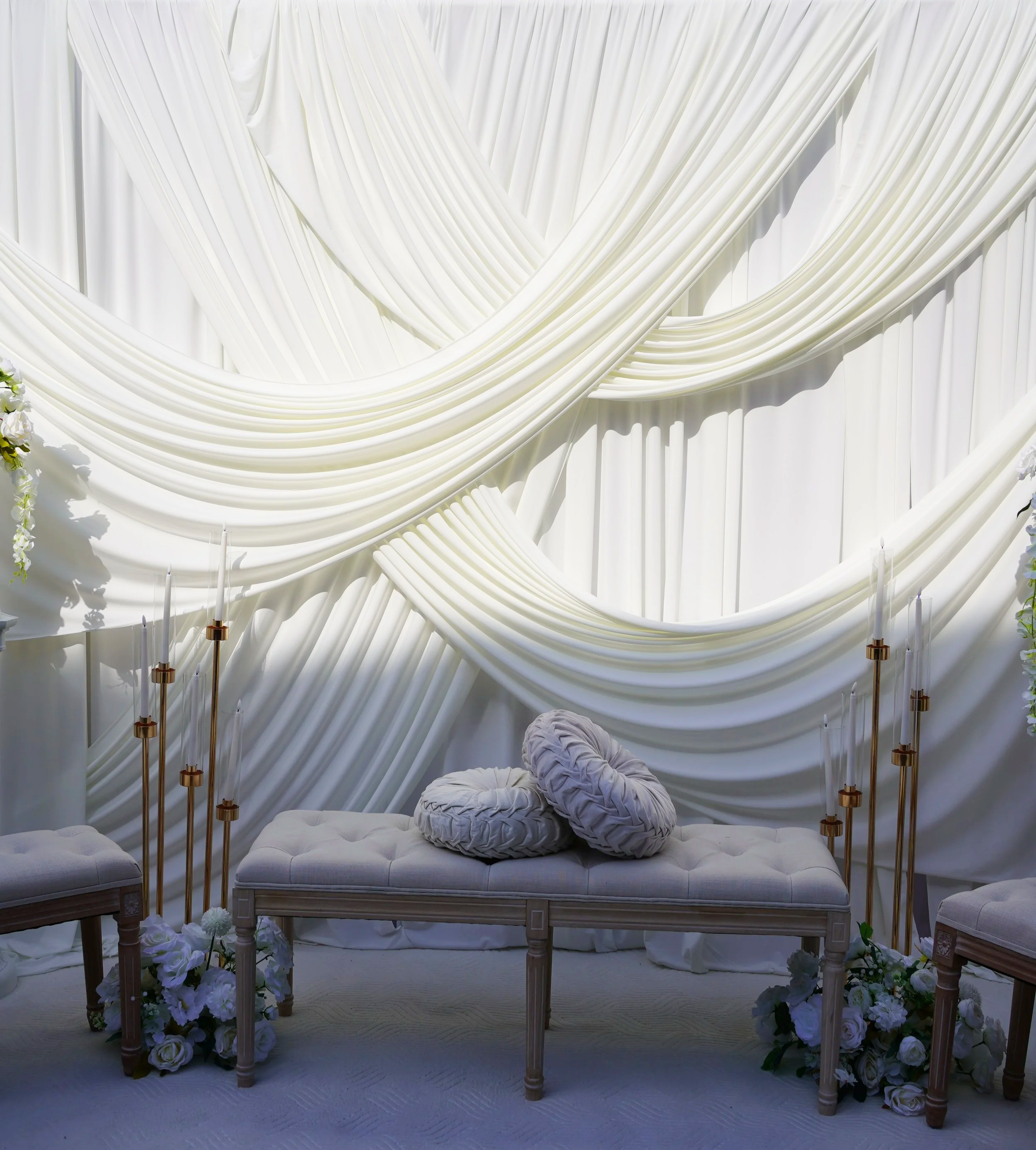 White draped fabric backdrop with gold candle holders and floral arrangements around a cushioned seating area, likely for a wedding or event.