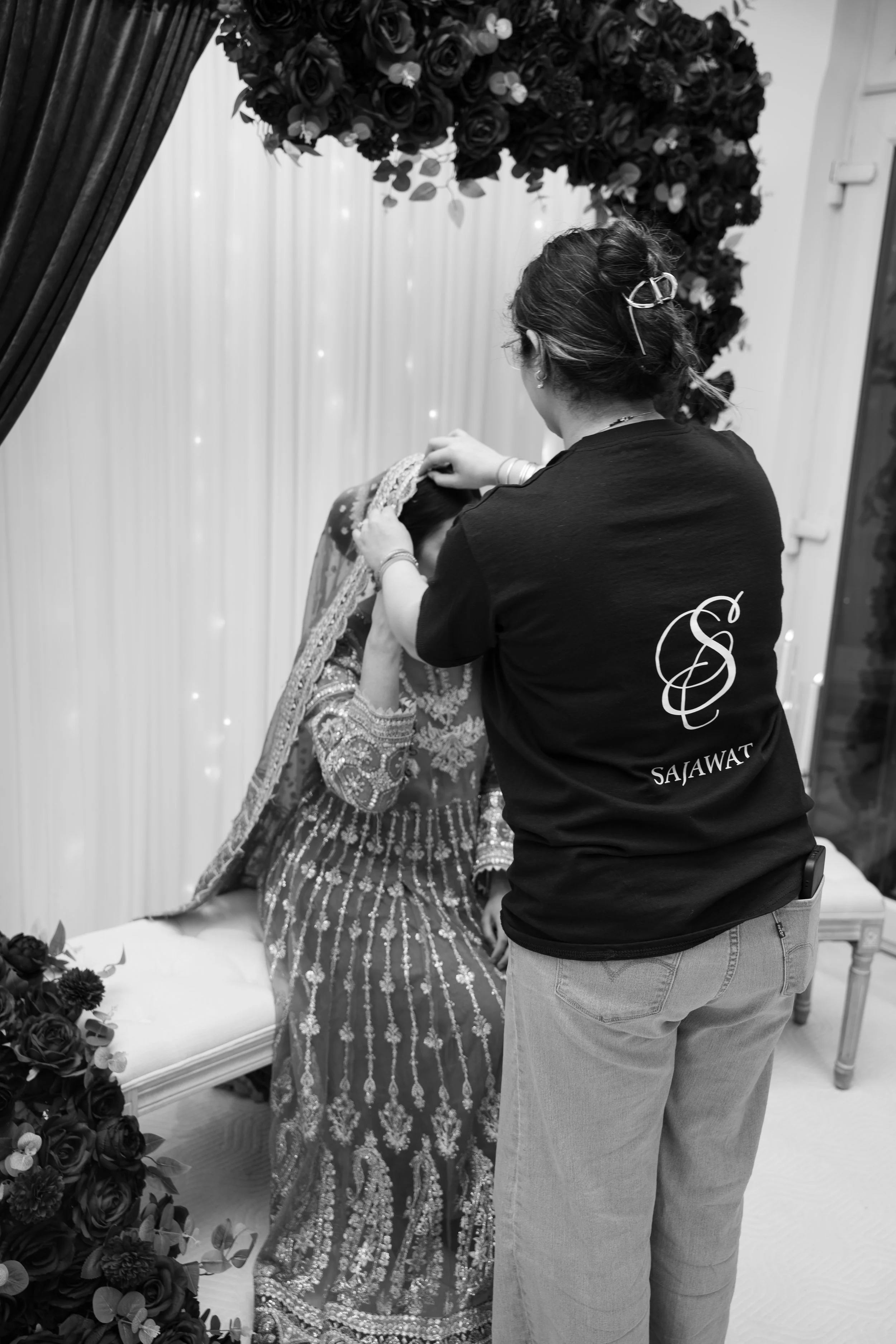 A woman dressed in traditional attire is seated while another woman stands behind her fixing a headpiece, in a decorated indoor setting with floral arrangements and draped backdrop.