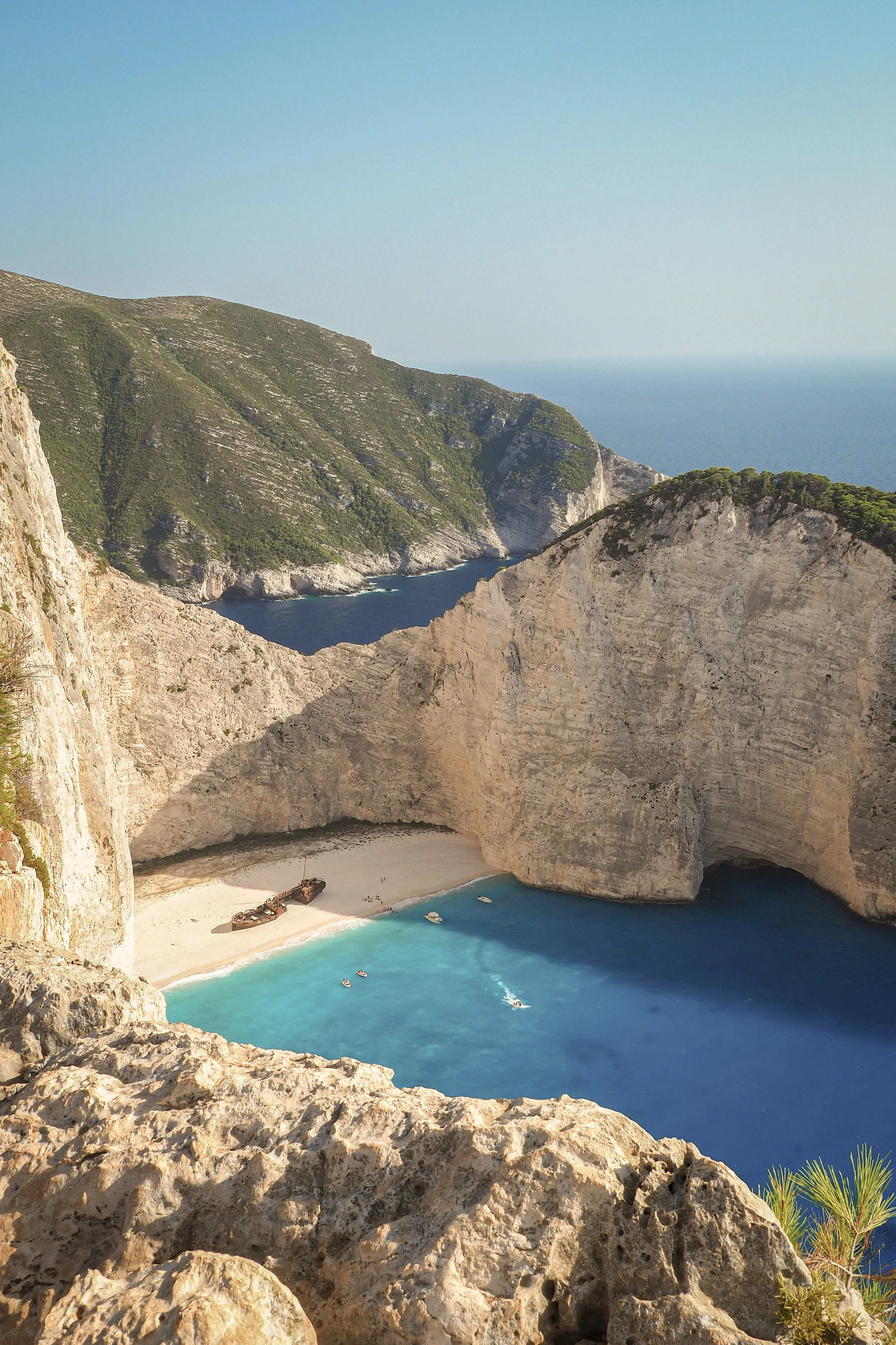 Crique de plage entourée de hautes falaises avec une mer turquoise, quelques bateaux et un bateau plus grand échoué sur la plage.