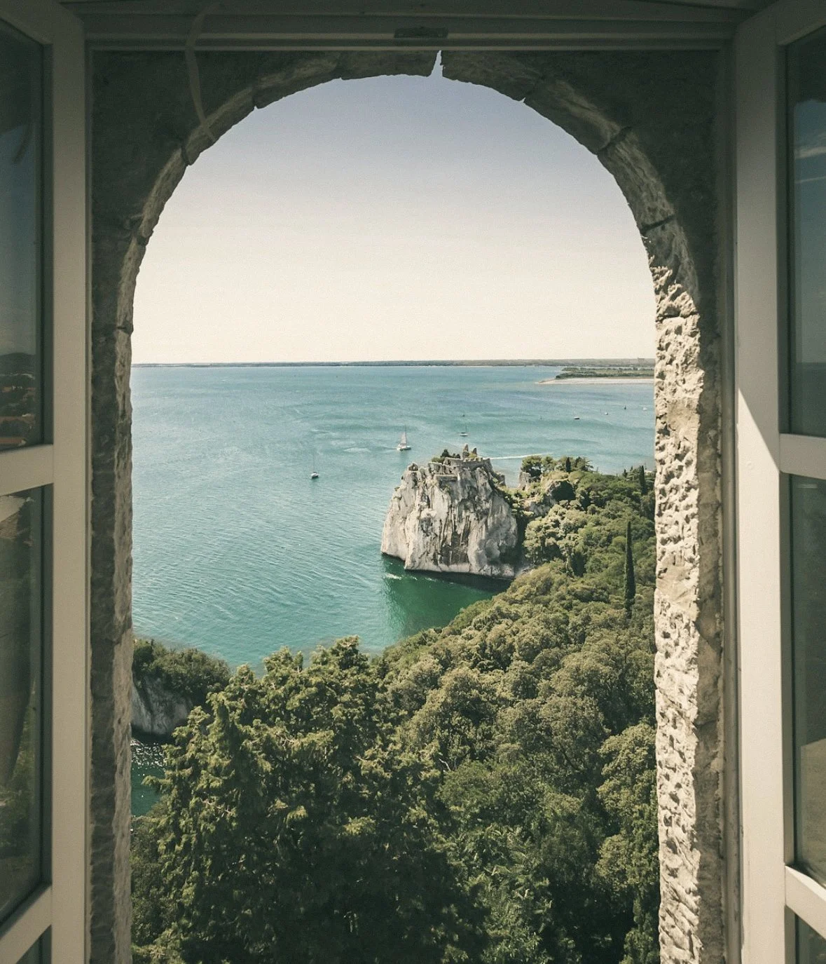 Vue d'une mer turquoise vue à travers une ouverture en forme d'arc dans un mur ancien en pierre, avec une formation rocheuse au centre entourée de végétation dense, et plusieurs voiliers en mer.
