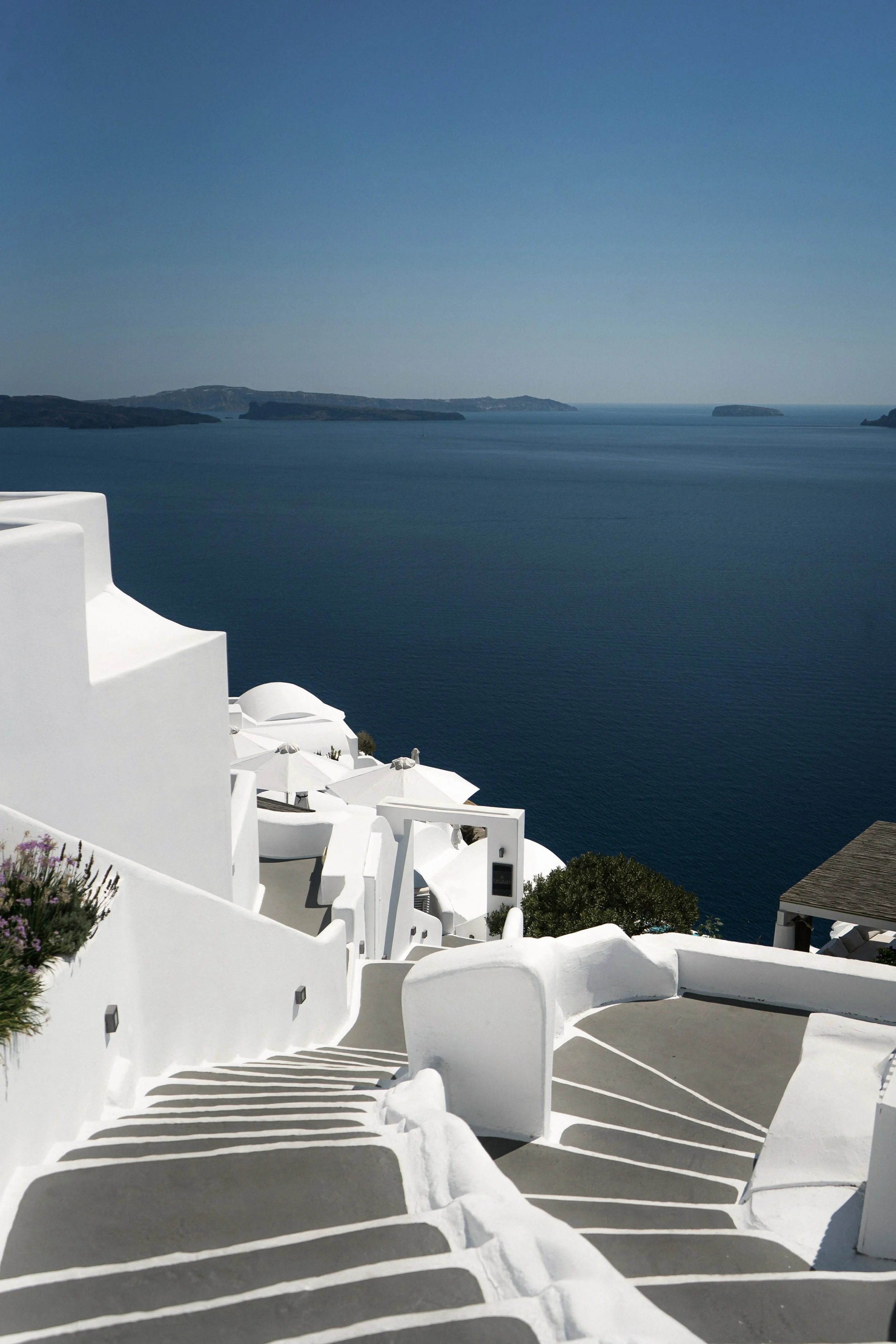 Vue de bâtiments blancs traditionnels sur une falaise en Grèce, avec la mer bleue et des îles à l'horizon, sous un ciel clair.