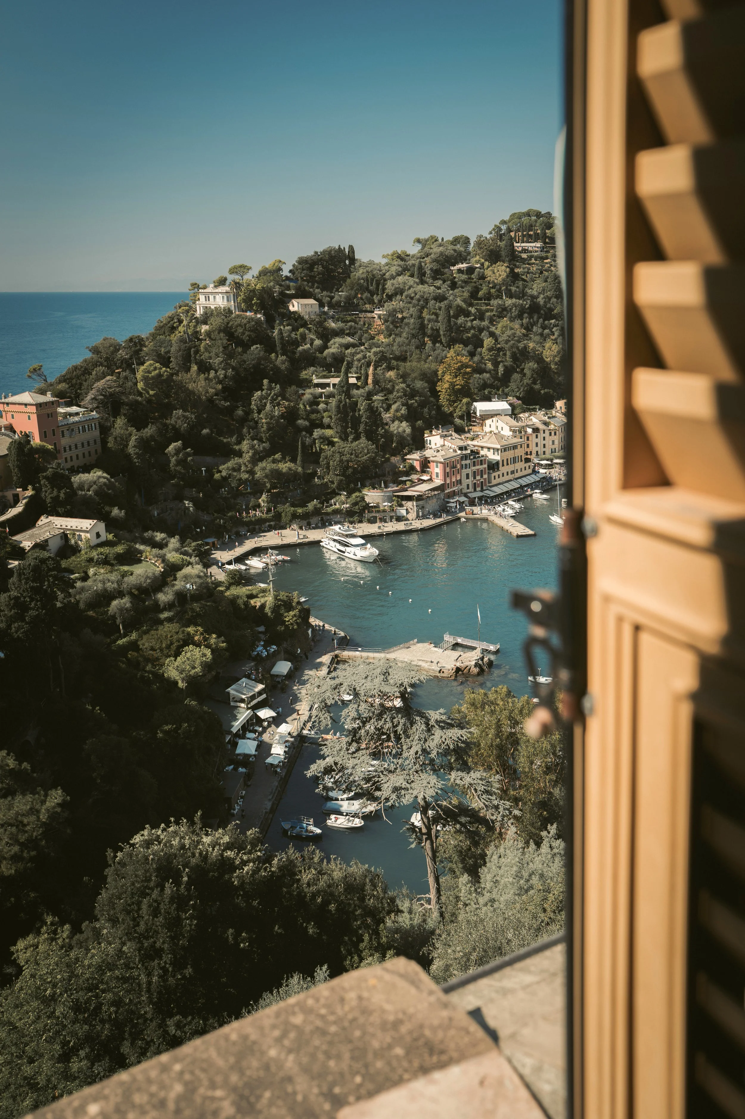 Vue d'une ville en bord de mer avec des bâtiments colorés, des bateaux dans le port, entourée de collines verdoyantes sous un ciel clair.