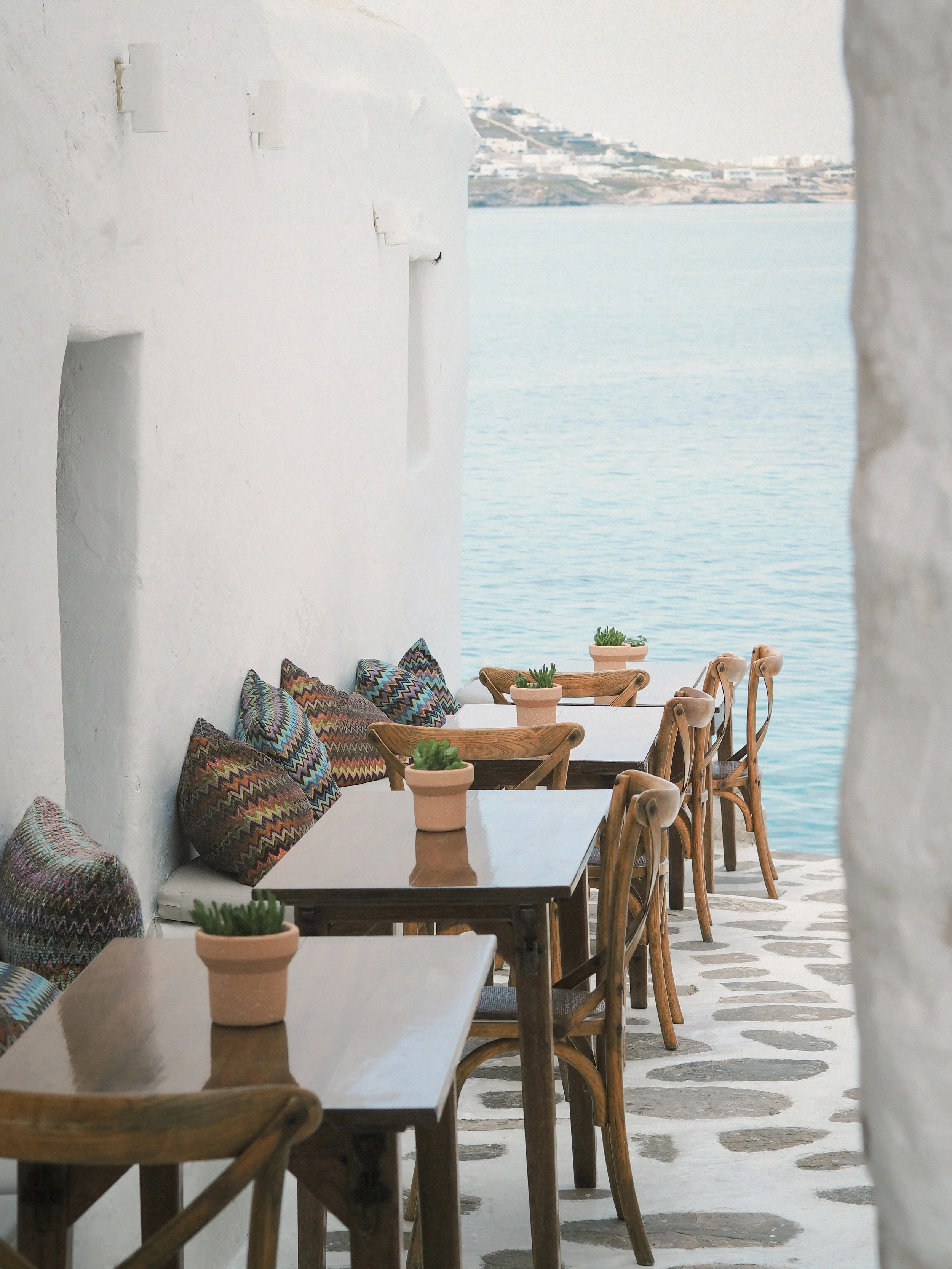 Terrasse avec tables en bois, chaises en bois, coussins colorés sur le banc, et petites plantes en pot, avec vue sur la mer et un paysage côtier en arrière-plan.
