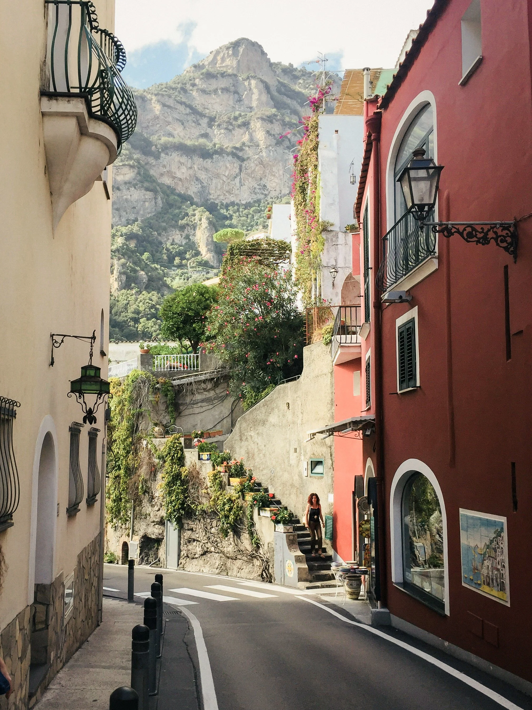 Rue étroite avec maisons colorées, plantes et montagnes en arrière-plan, un femme marche sur la rue.