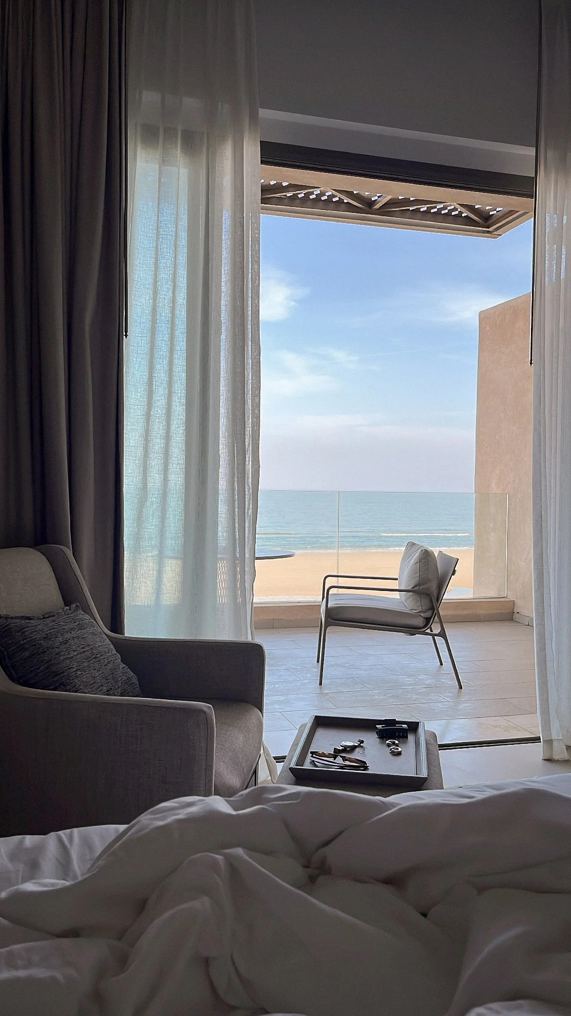 Vue depuis un lit d'une pièce d'hôtel, avec une vue sur la mer, un balcon avec une chaise en plastique, rideaux transparents, et une partie du mobilier intérieur visible.