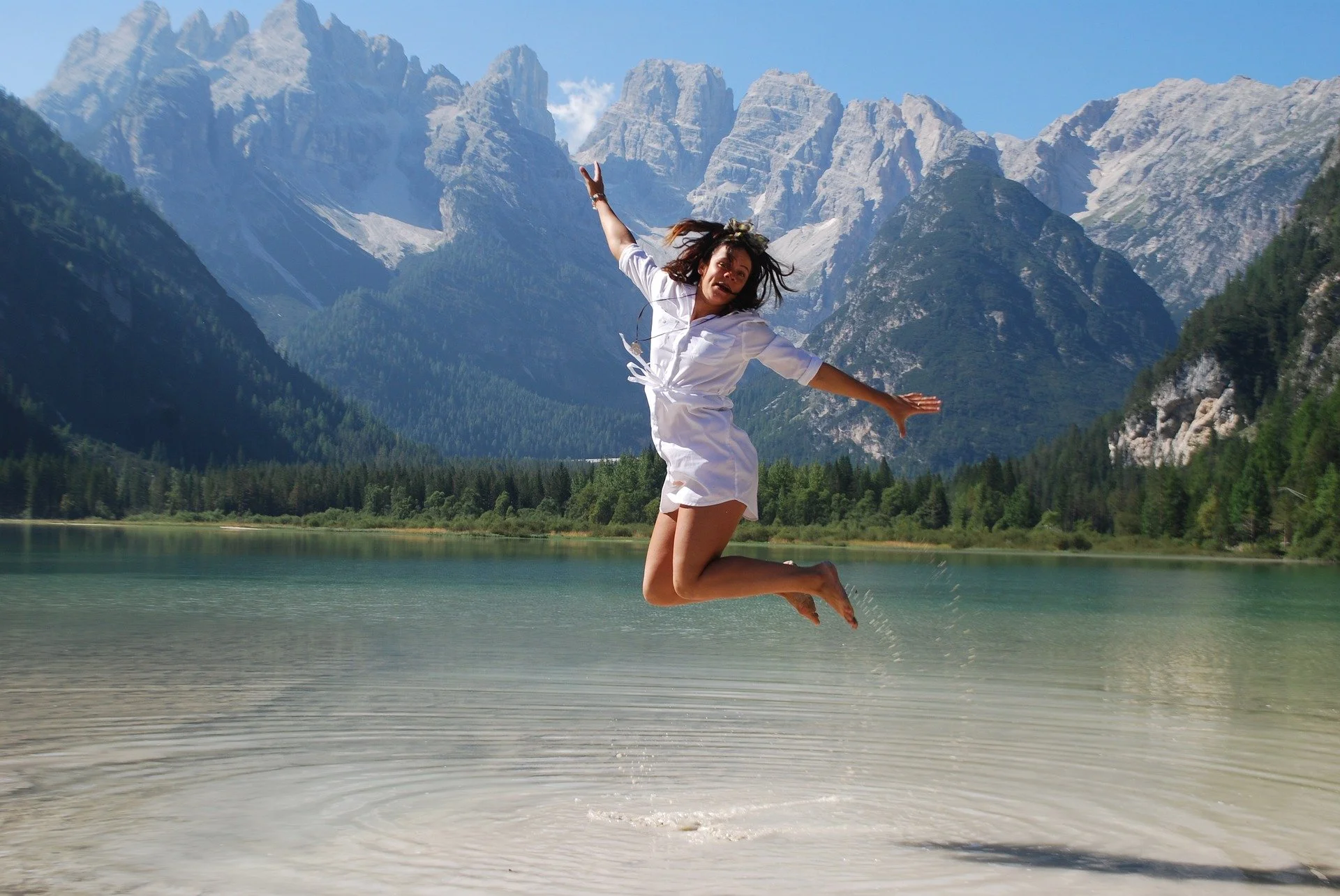 A woman in a white dress jumping above the water of a lake with a mountainous landscape in the background.