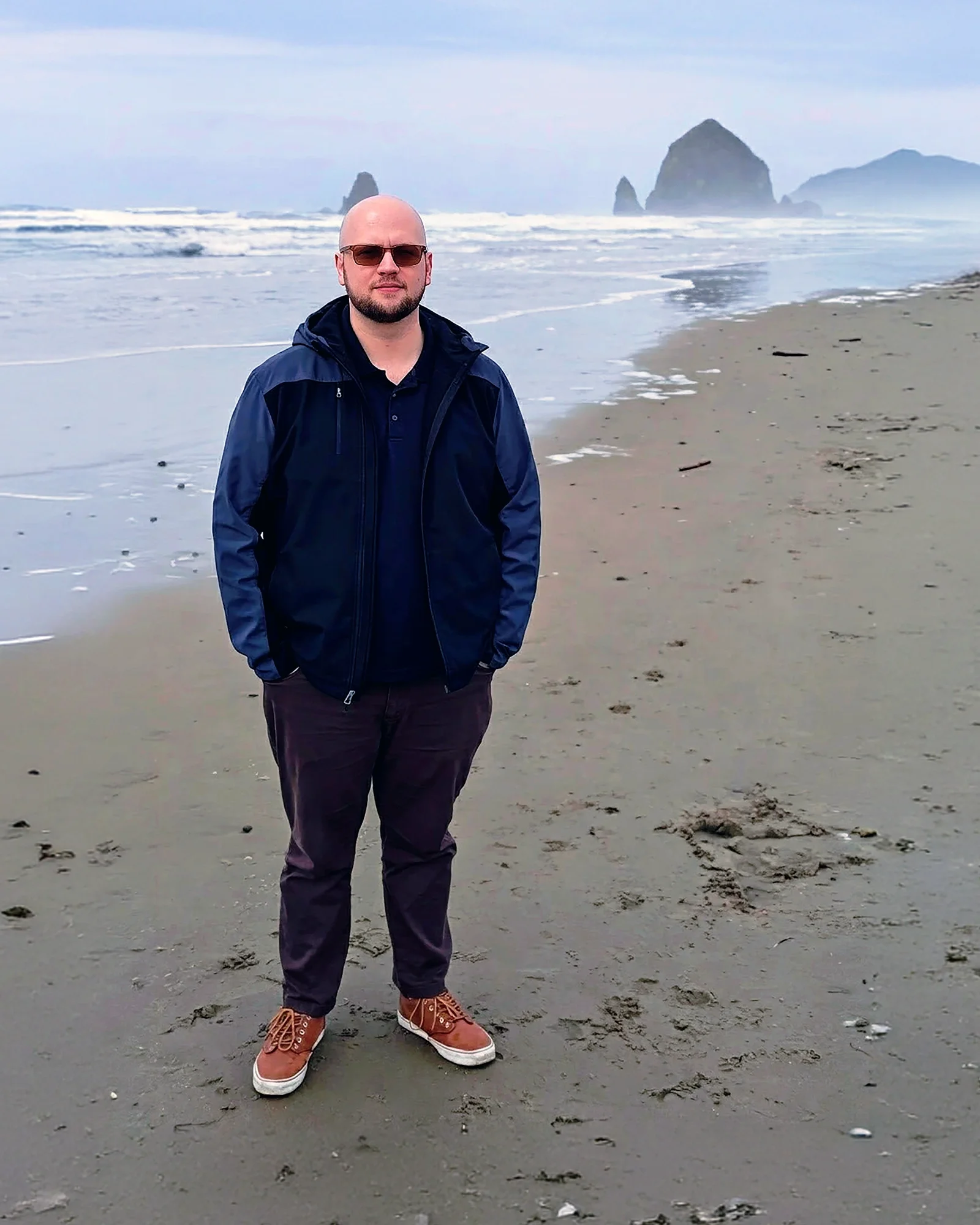 Kenton Summers standing on a beach with large rock formations in the ocean in the background, wearing sunglasses, a dark rain jacket, dark pants, and orange sneakers.