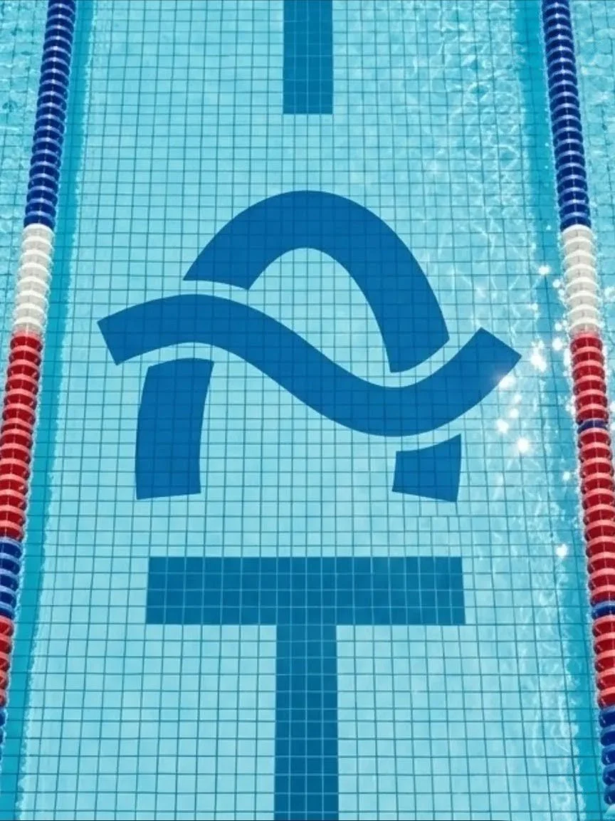 A swimming pool with tiles and lane markers in blue, white, and red, with a large logo of a stylized letter 'R' and a wave pattern painted on the pool floor.