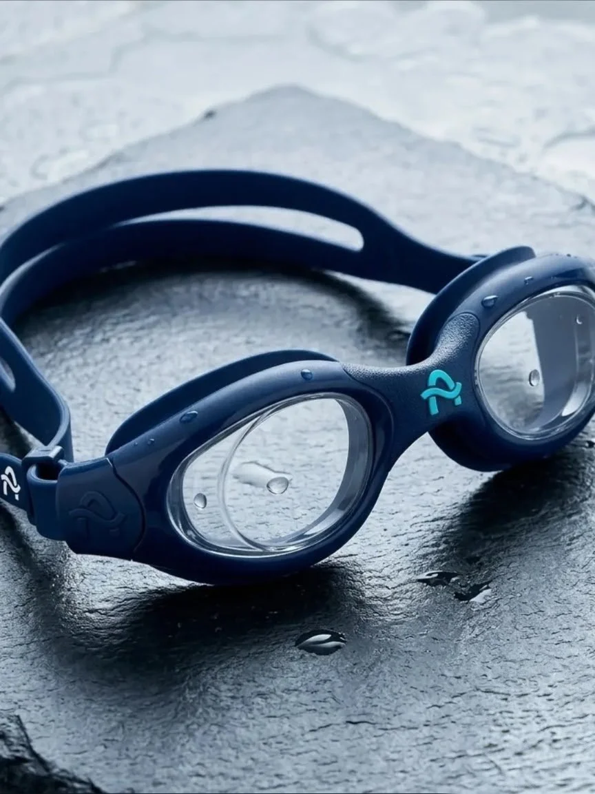 Close-up of black swimming goggles with water droplets, placed on a wet, textured black surface.